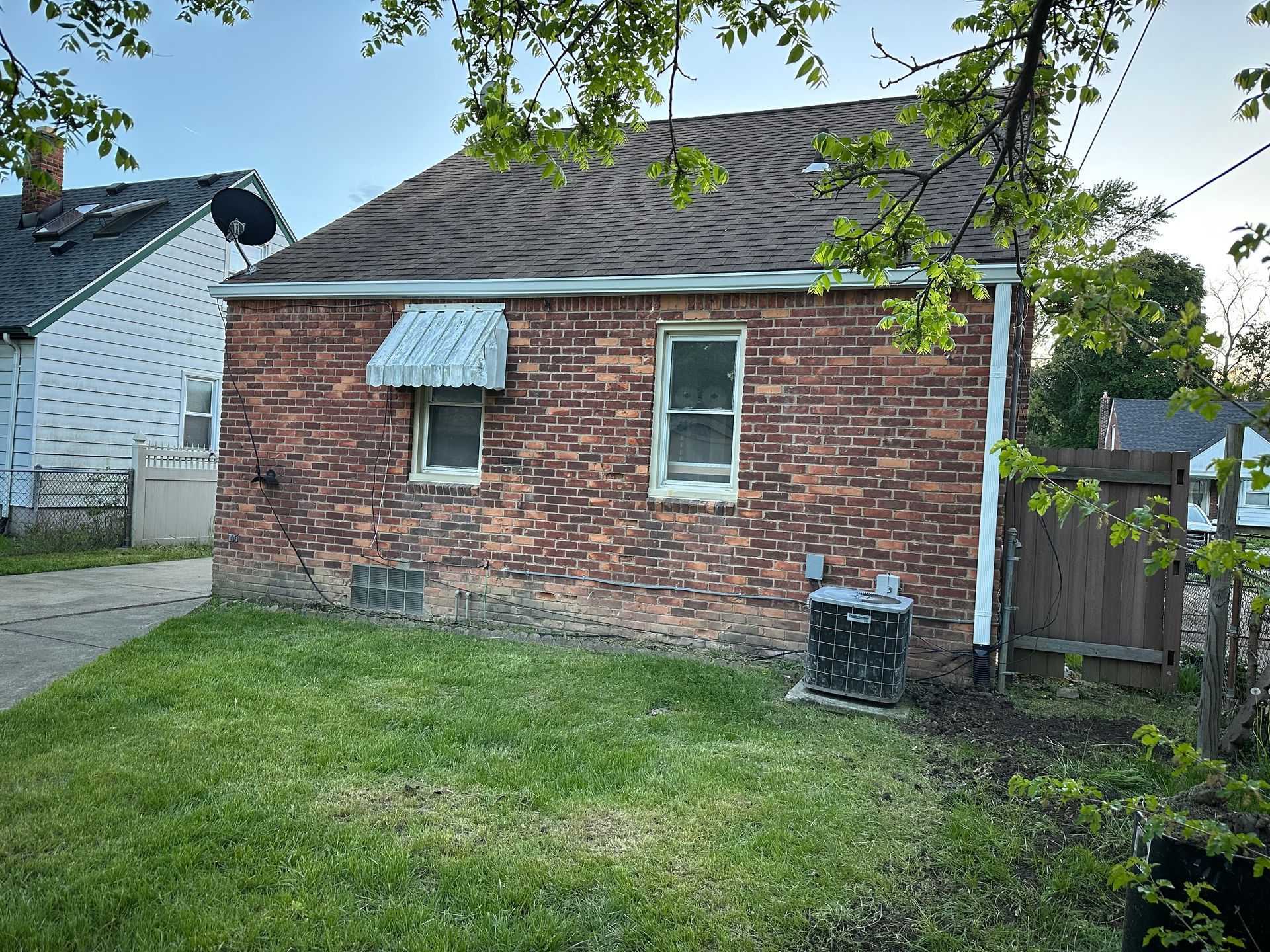Back of a small brick house with a weathered roof, two windows, and an awning over one. Green grass in the foreground.