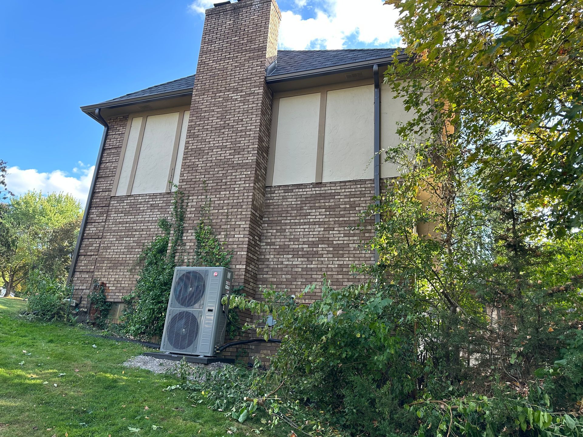 Brick house with chimney, HVAC unit, and overgrown landscaping.