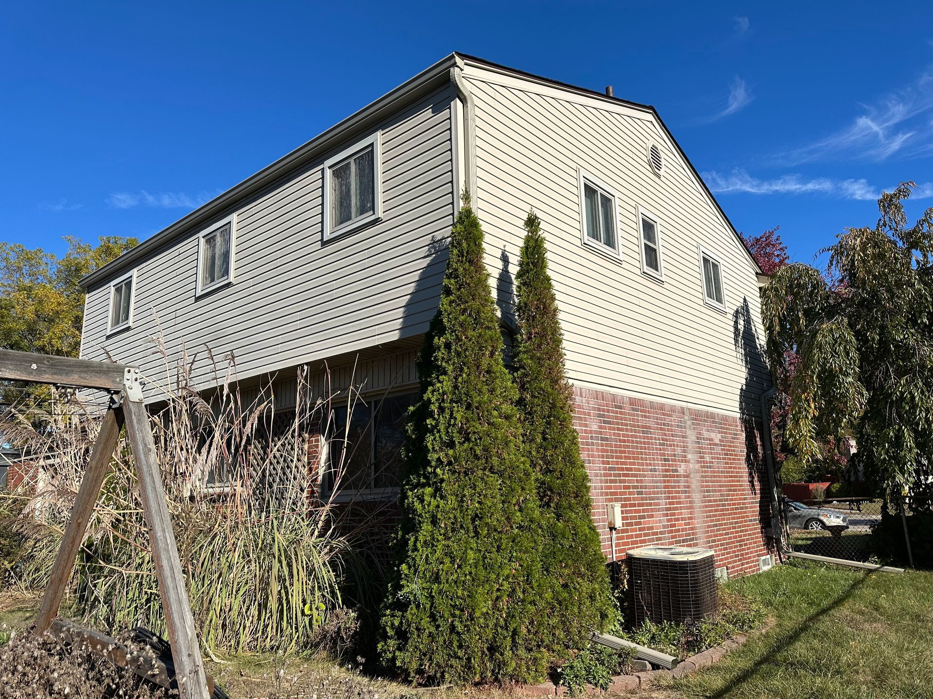 Two-story house with light siding and brick base. Green trees in front, blue sky.