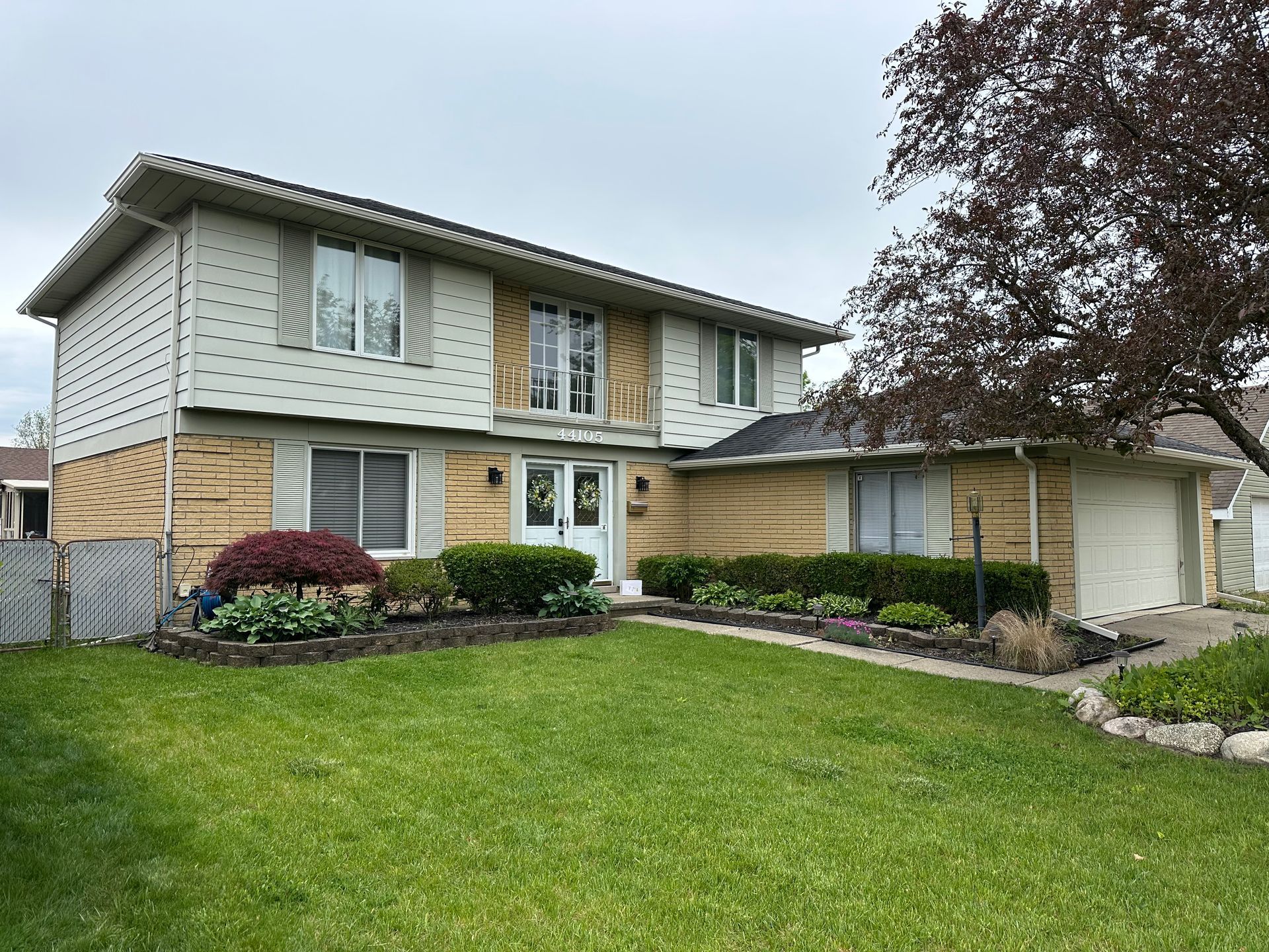 Two-story house with tan brick and beige siding. Green lawn and landscaping. Cloudy sky.