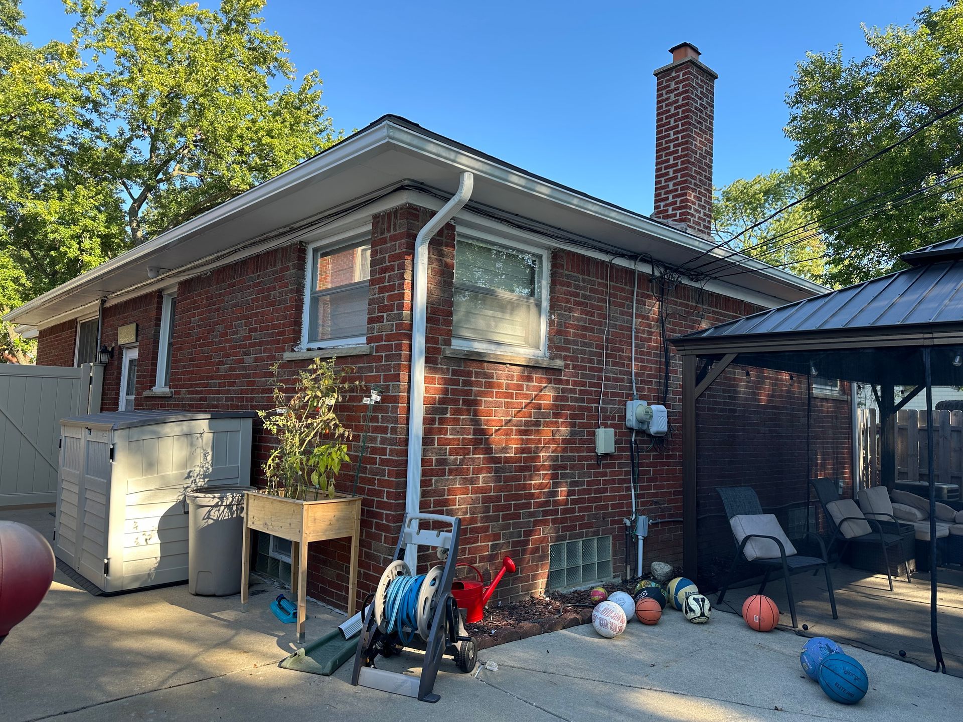 Brick house exterior with white trim, downspouts, and a gazebo on a paved patio.