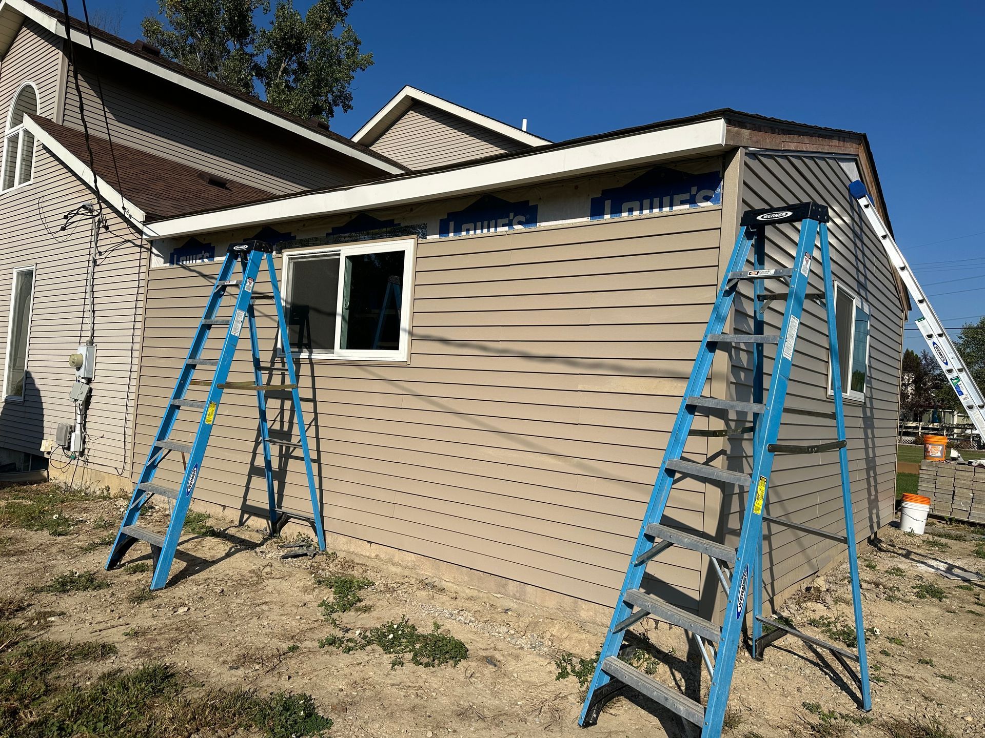 Building exterior with tan siding, blue ladders, and a white window against a blue sky.