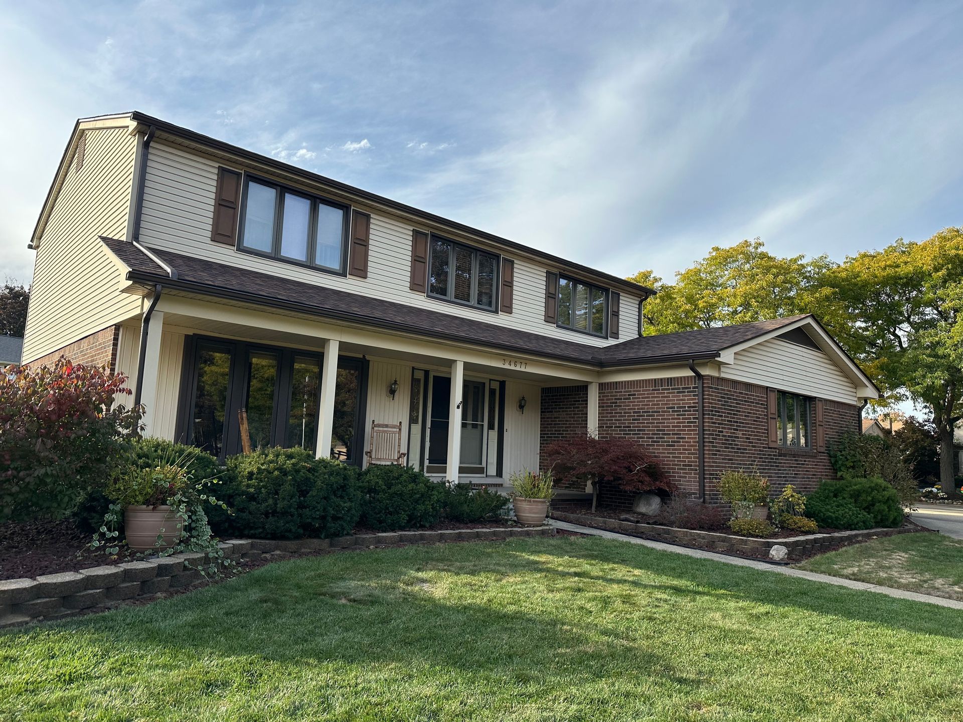 Two-story house with beige siding, brown roof and trim, and green lawn on a sunny day.