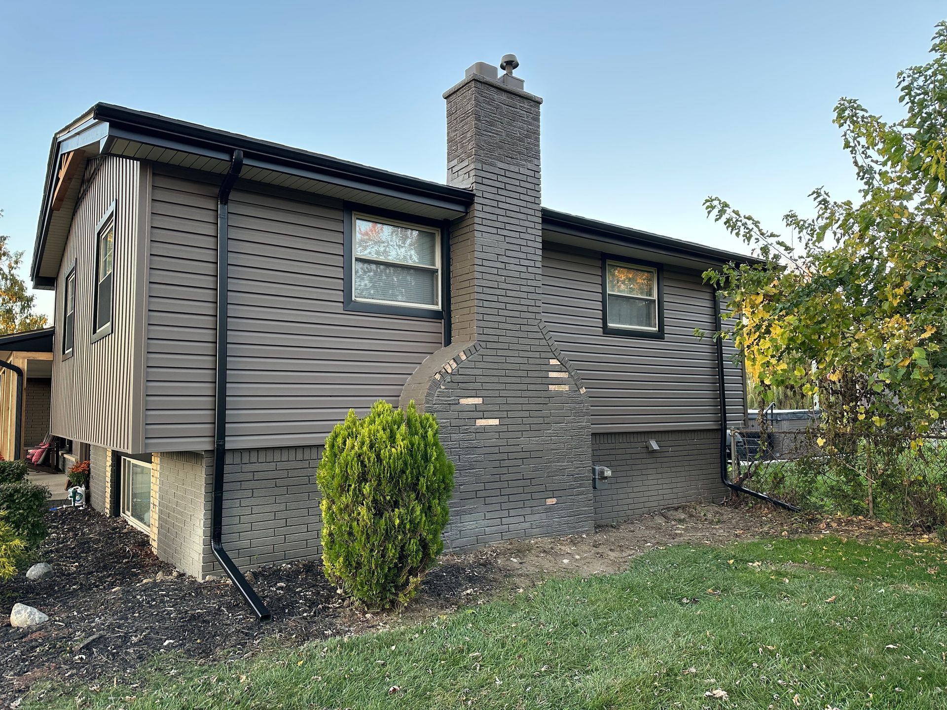 Two-story house with gray siding, brick chimney, windows, black gutters, and a small green bush.