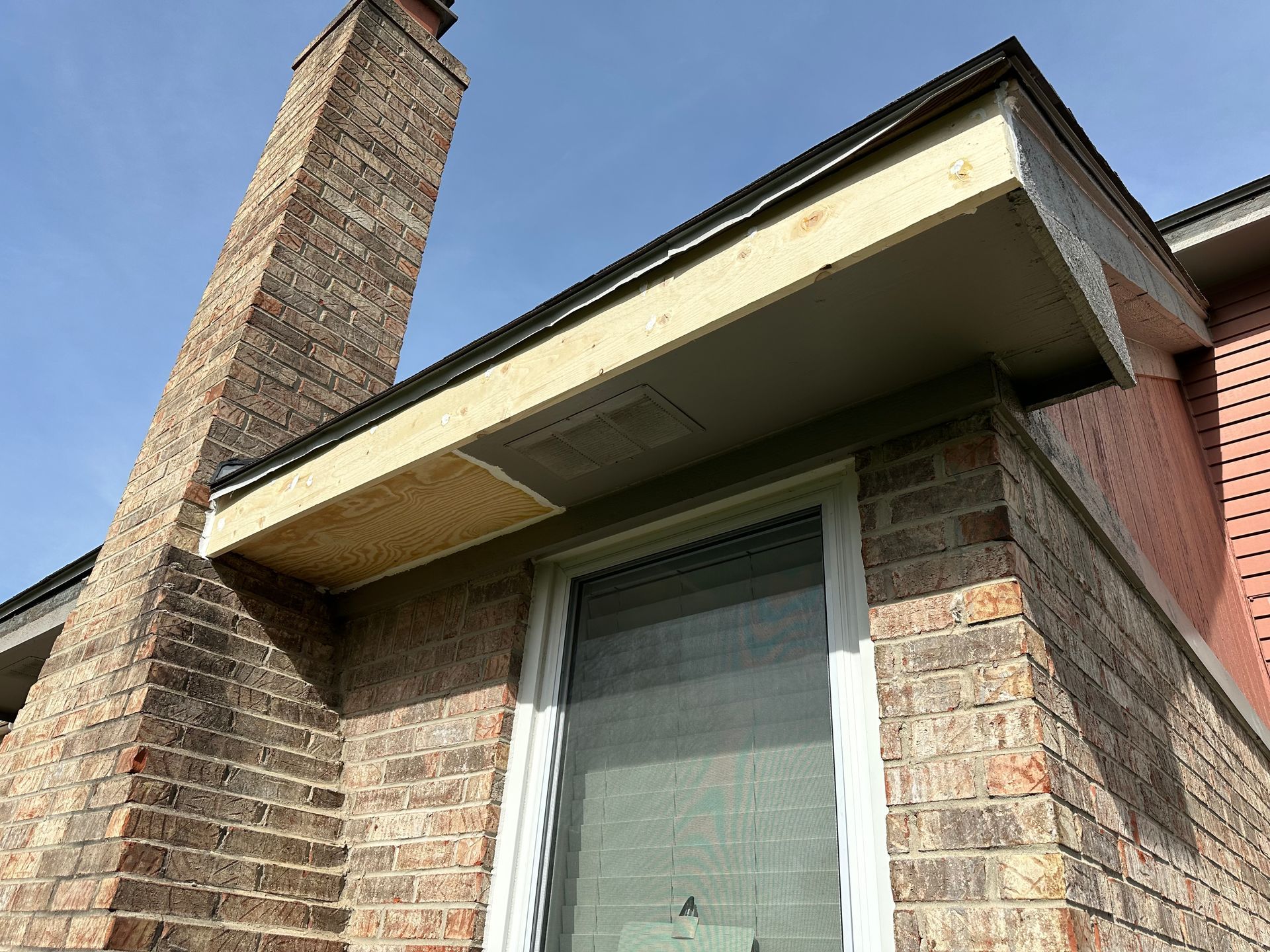 Brick building exterior with chimney, window, and overhanging roof under a blue sky.