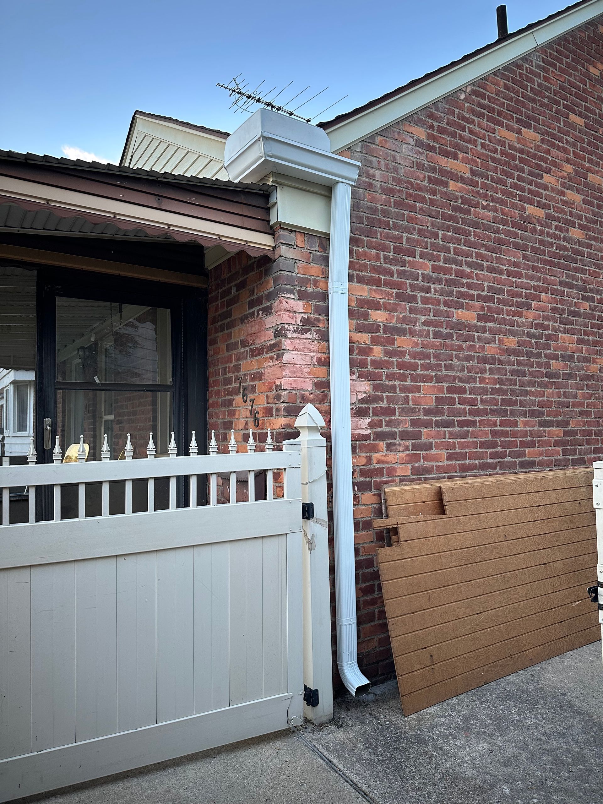 Brick building with a white gutter, downspout, and fence. A pegboard leans against the brick.