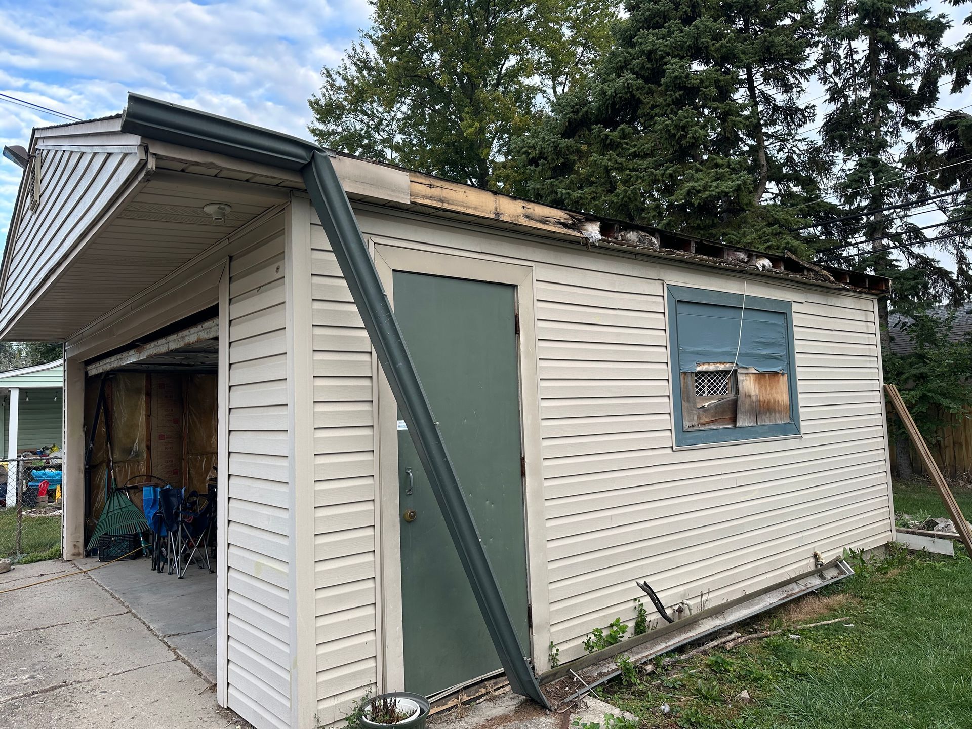 Garage with damaged roof, green door and trim, tan siding, and broken window.