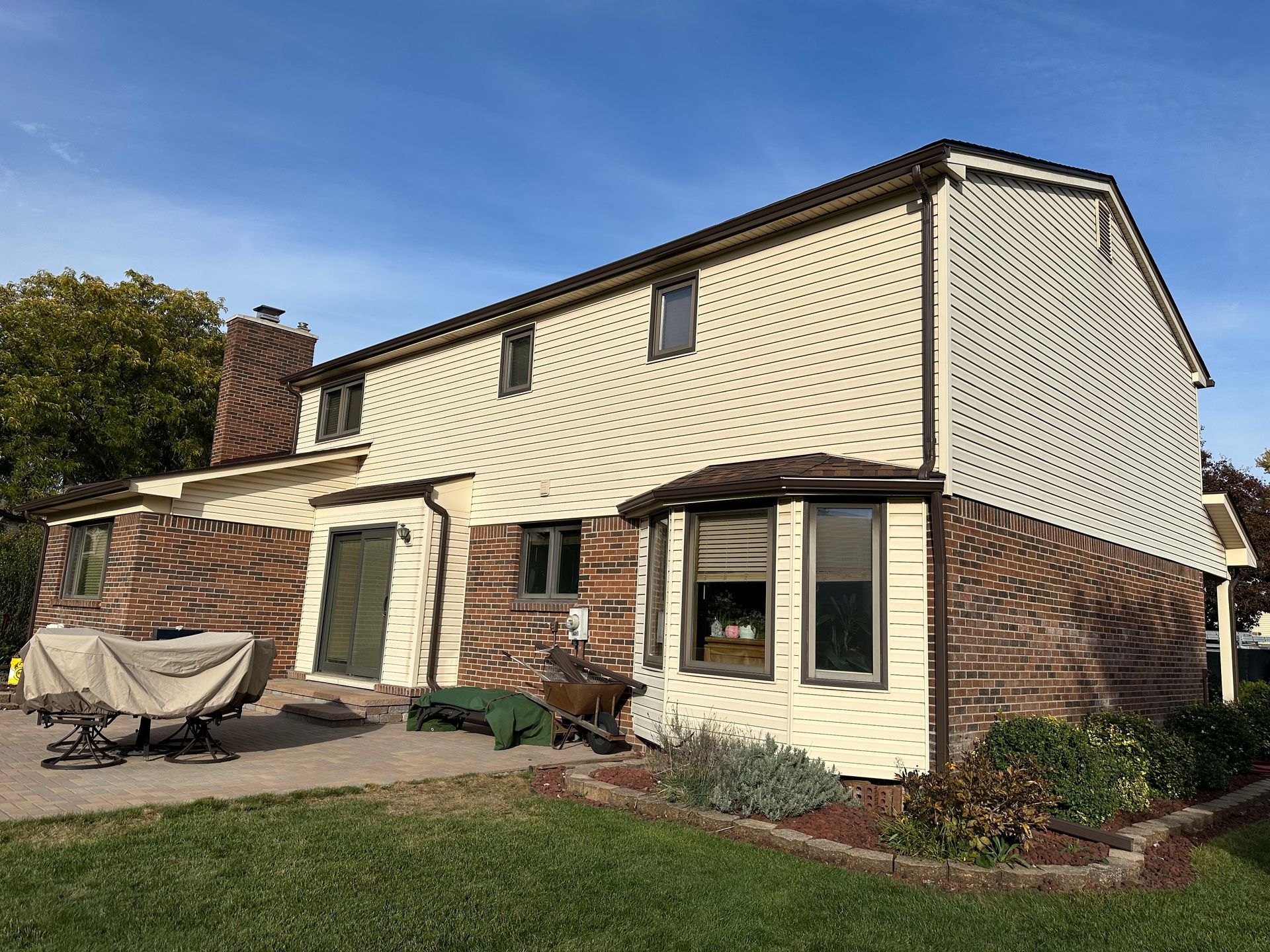 Two-story beige house with brown trim and brick siding under a blue sky. Backyard patio with grass.