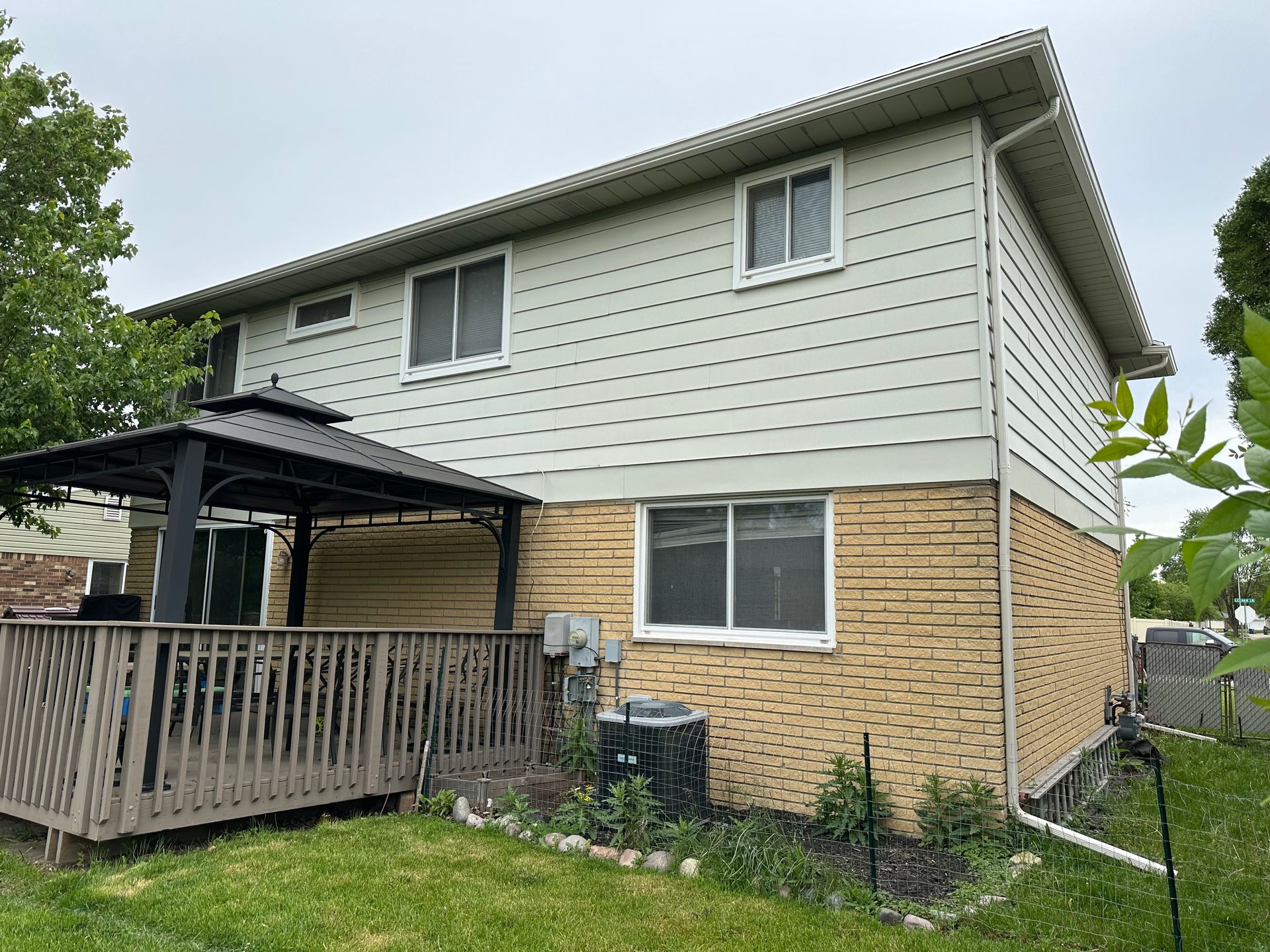 Two-story house with tan siding and brick base, deck, and gazebo on a cloudy day.