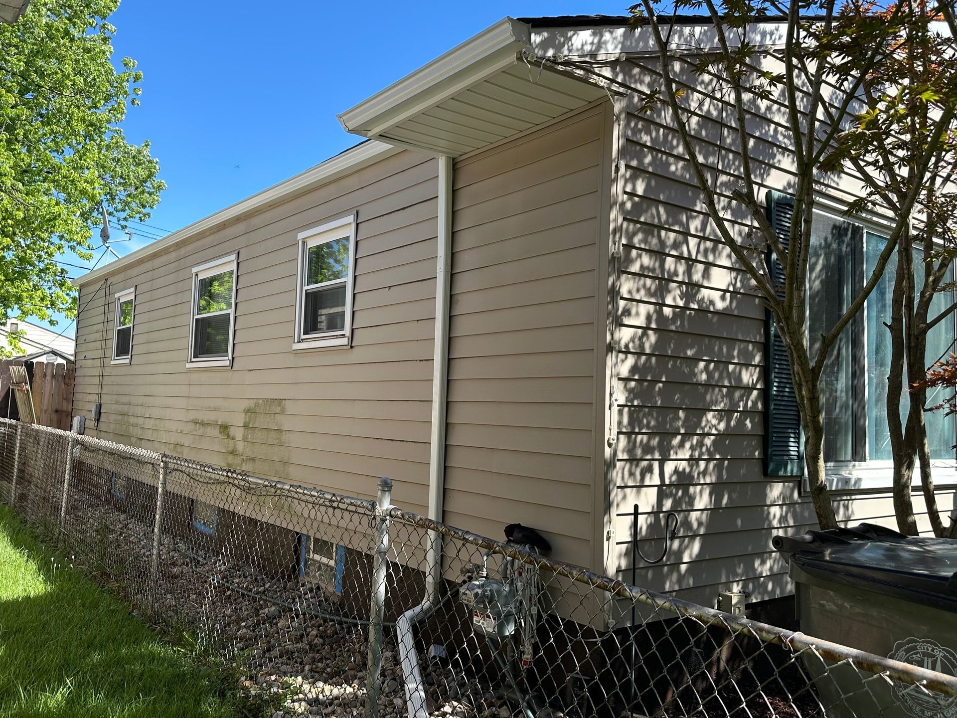 Tan-sided building with windows and white trim next to a chain-link fence and green tree foliage.
