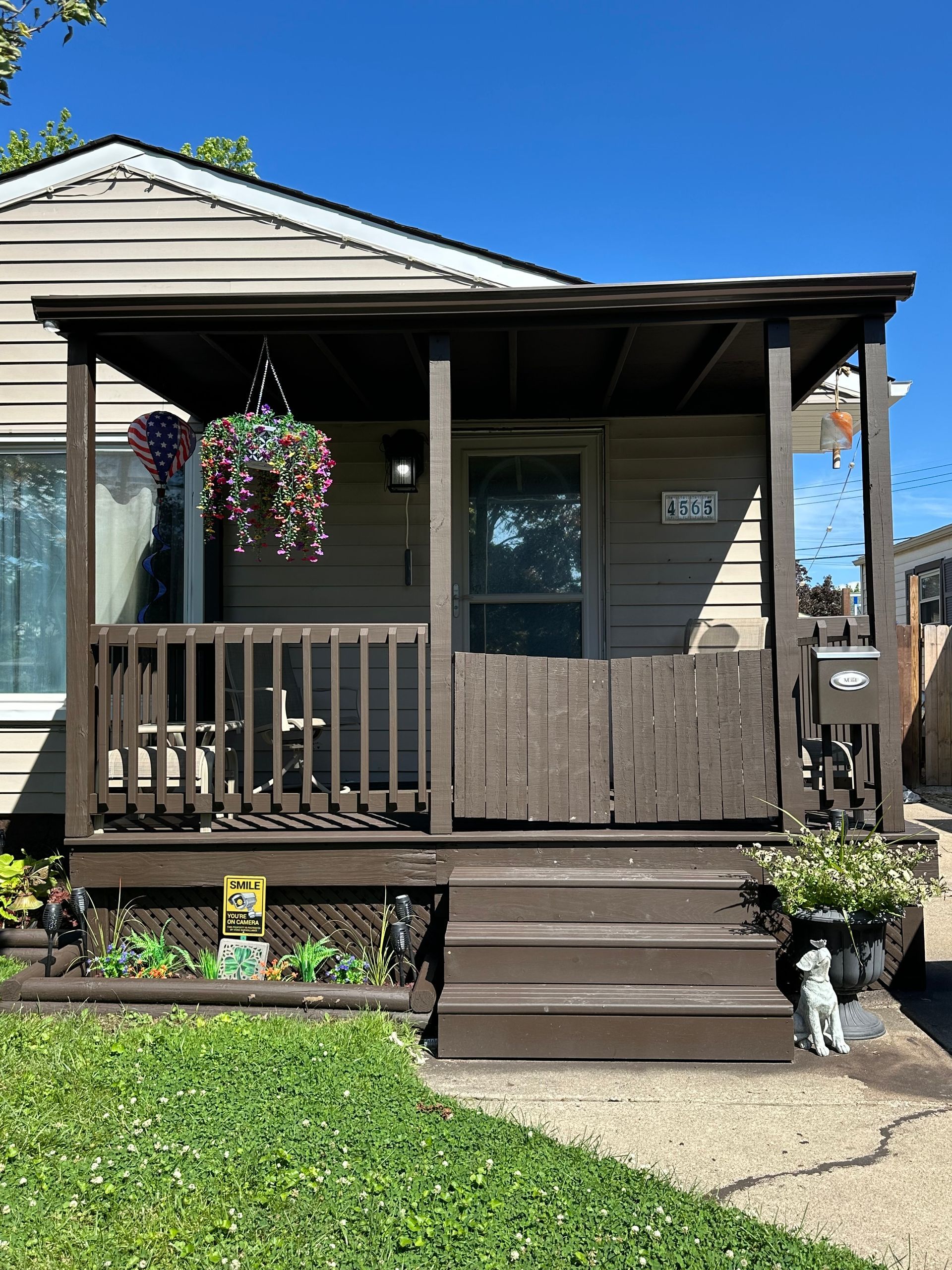 Brown porch with hanging plants, steps, and a dog statue in front of a tan house.