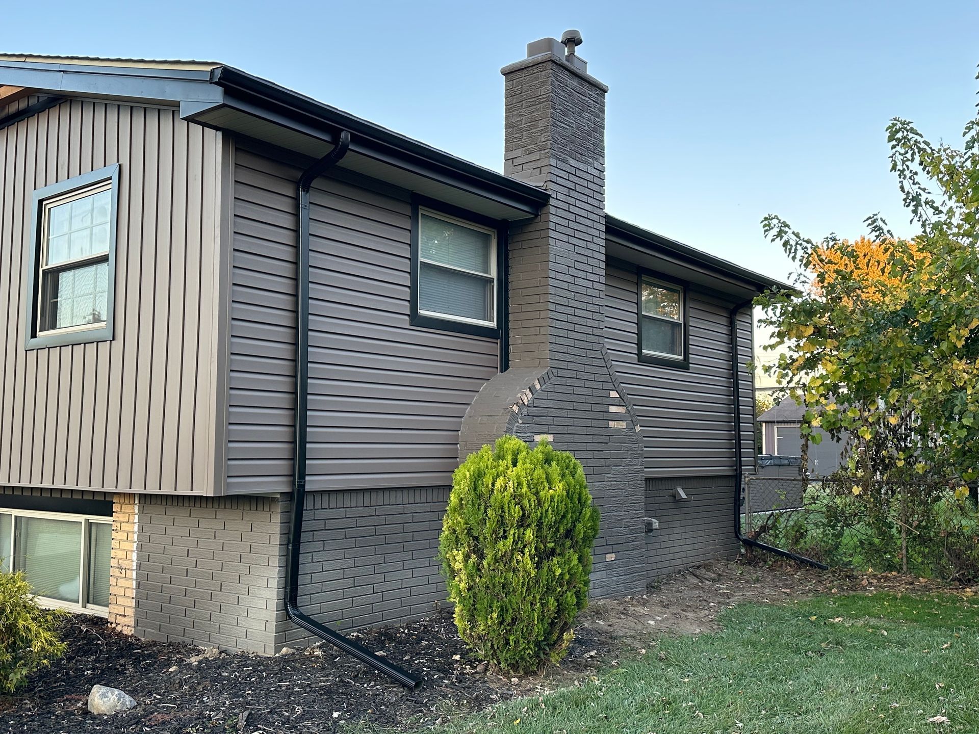 Side view of a house with gray siding, black trim, and a brick chimney. A small green bush is in front.