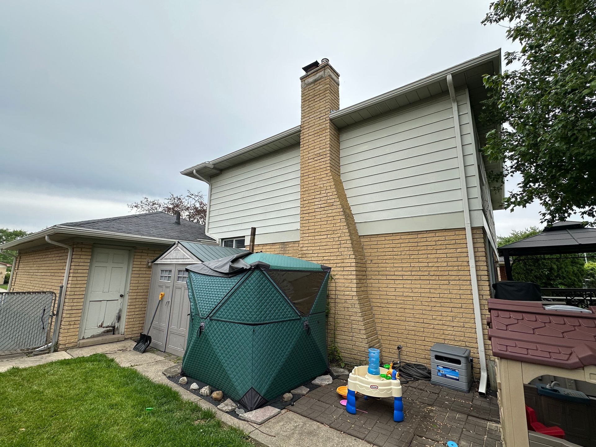 Backyard with a brick house, chimney, garage, green play tent, and play table under a cloudy sky.