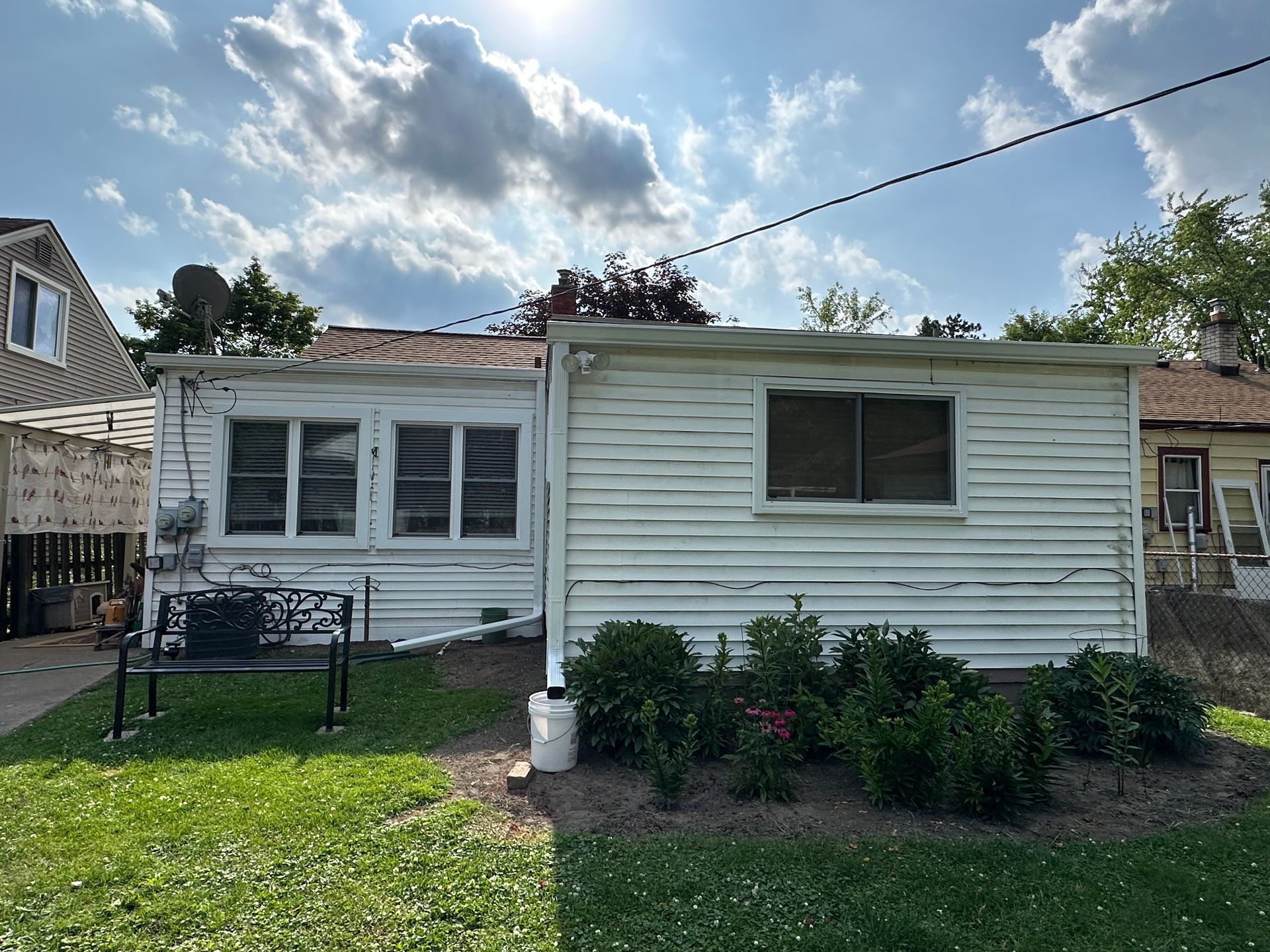 White, weathered, one-story house with two windows and a small garden, under a partly cloudy sky.