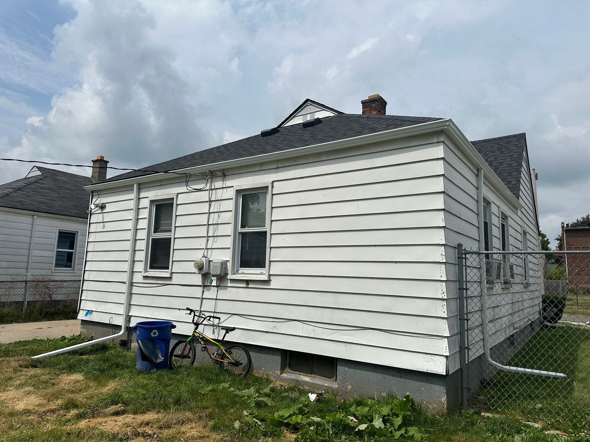White house with a dark roof, windows, and chain link fence. Green grass and cloudy sky are in the background.