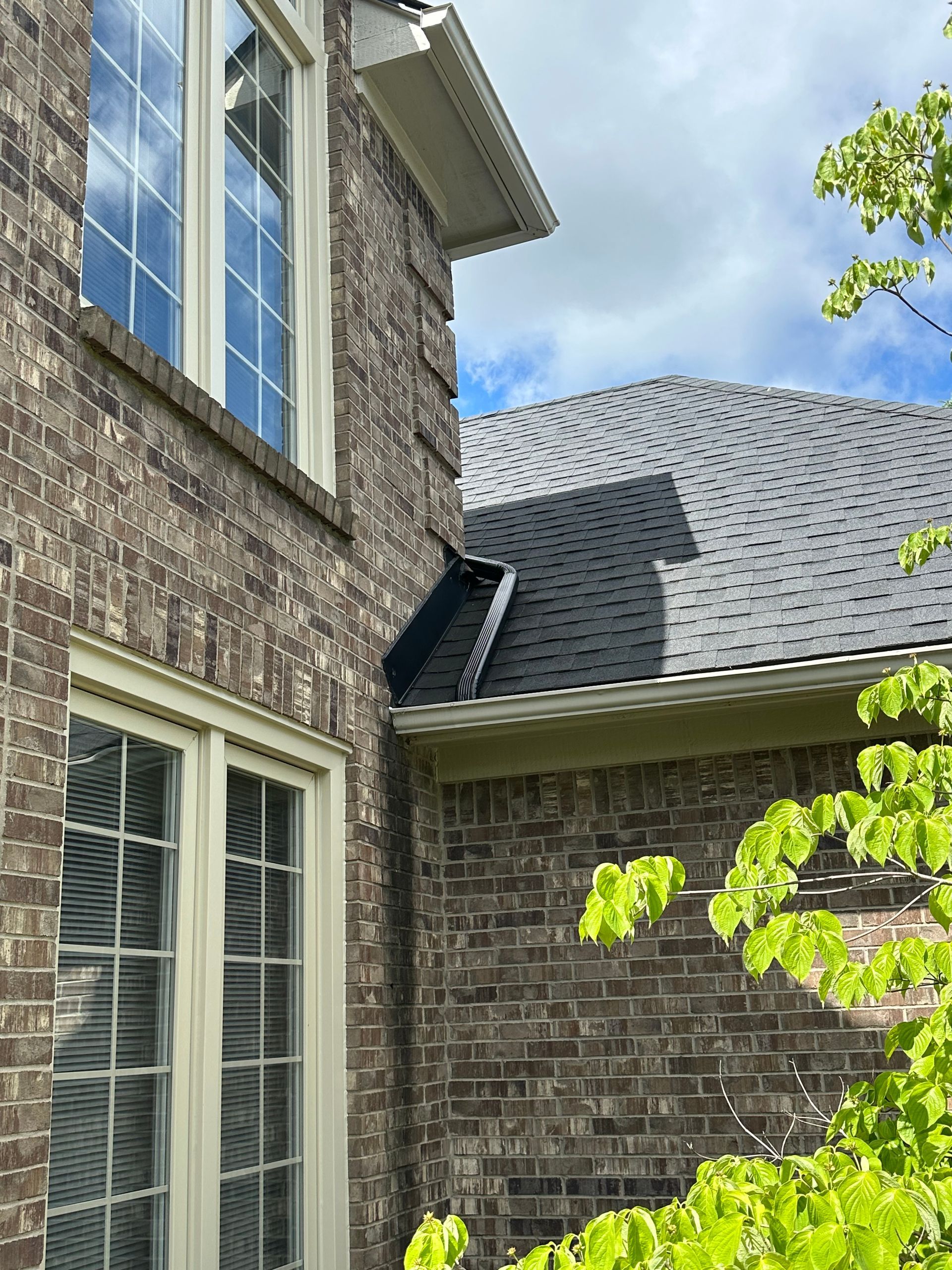 Brick house with windows; dark shingled roof, trees in the foreground, cloudy sky.