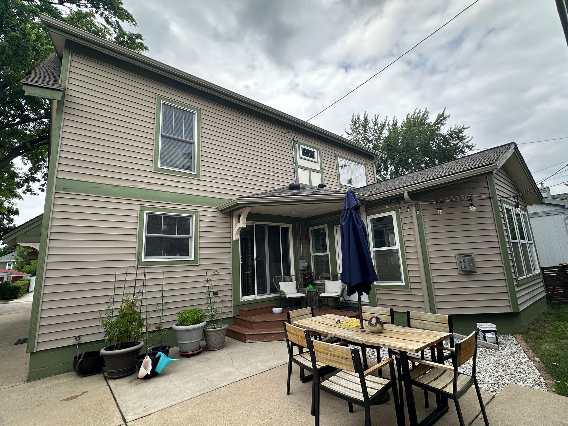 Back of a two-story beige house with a patio, outdoor dining set, and an umbrella, under a cloudy sky.