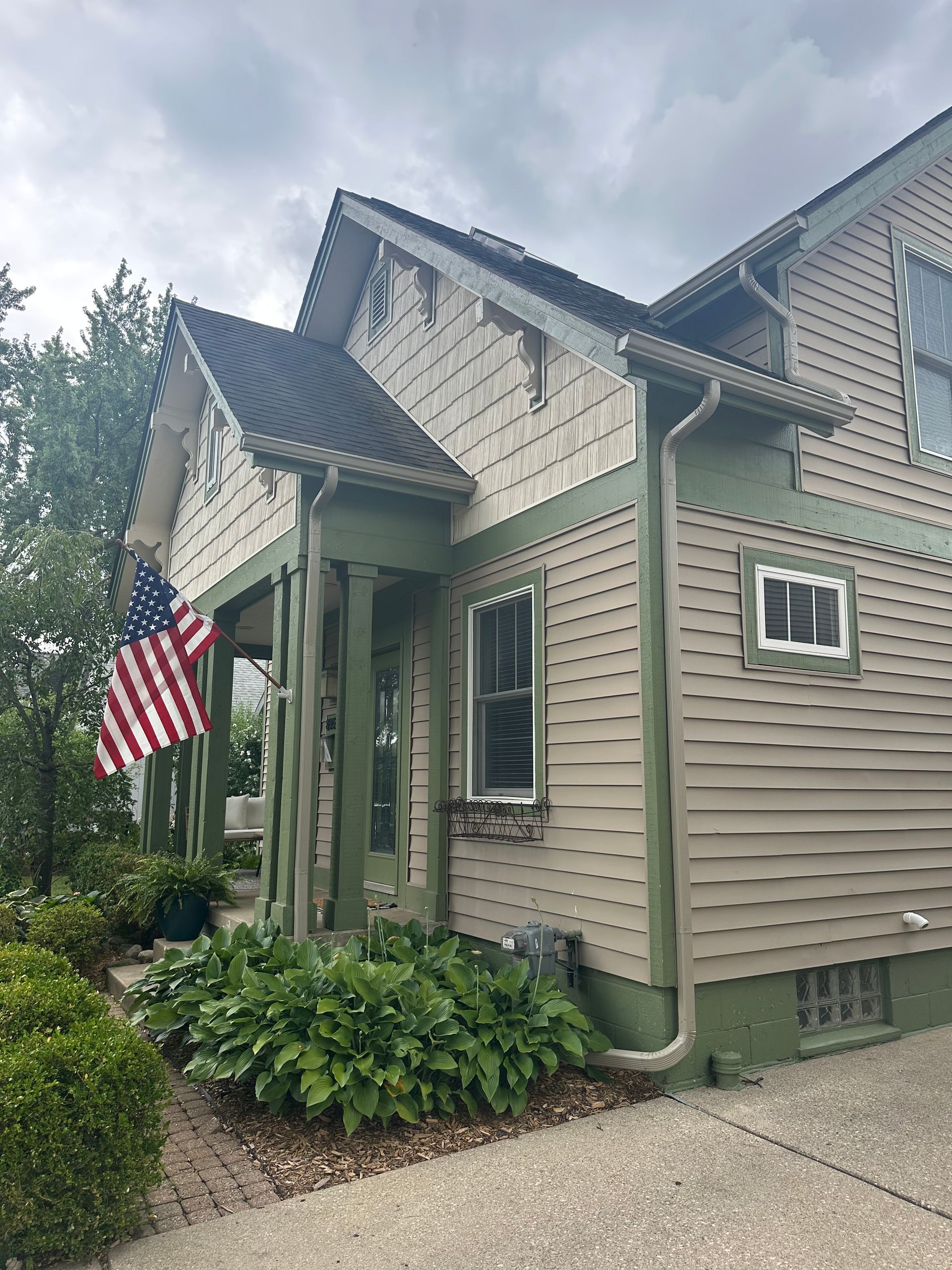 Two-story house with green trim and tan siding. American flag hangs on the front porch. Cloudy sky.