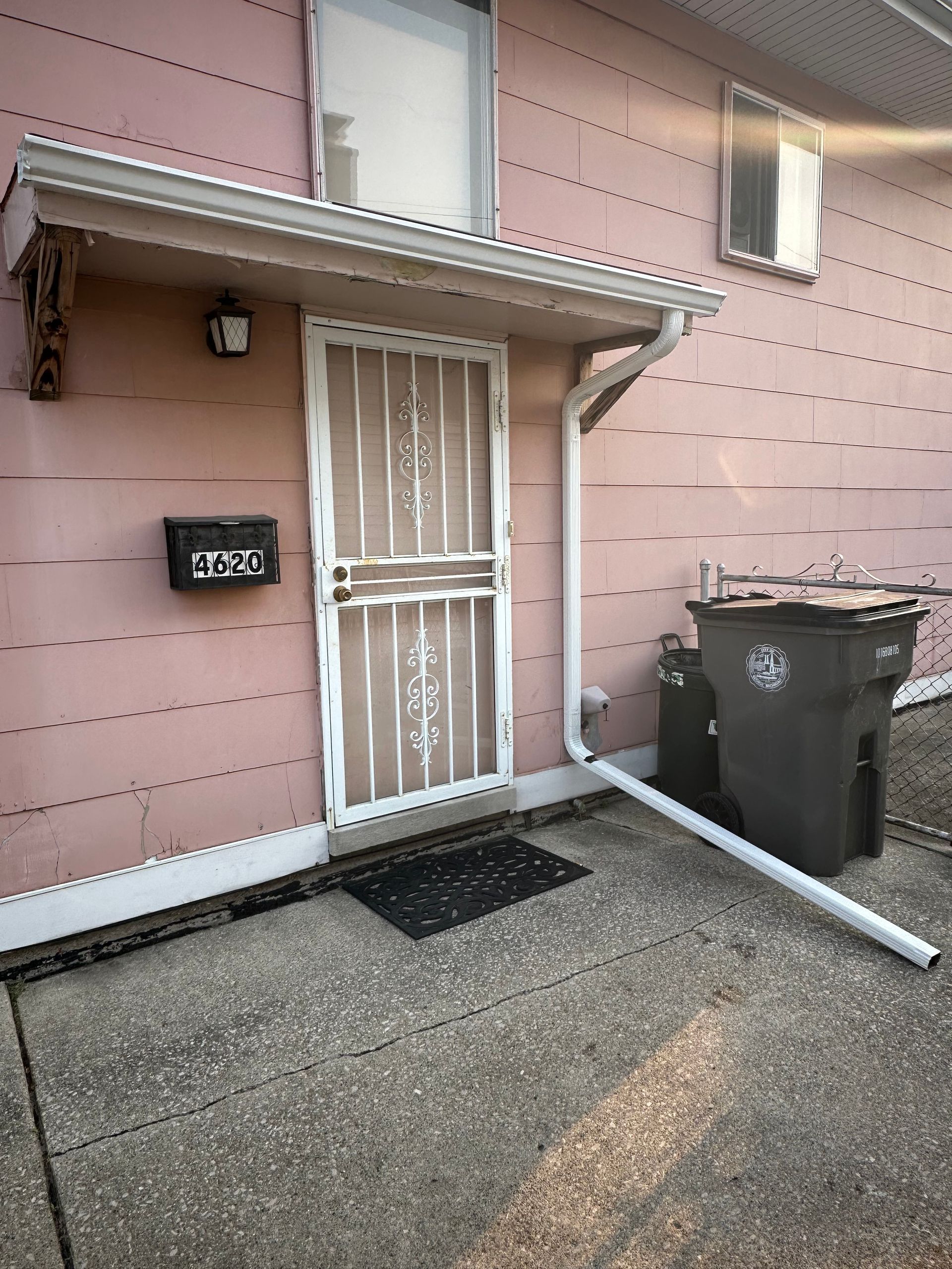 Pink house exterior with security door, awning, mailbox, and trash bins on a concrete patio.