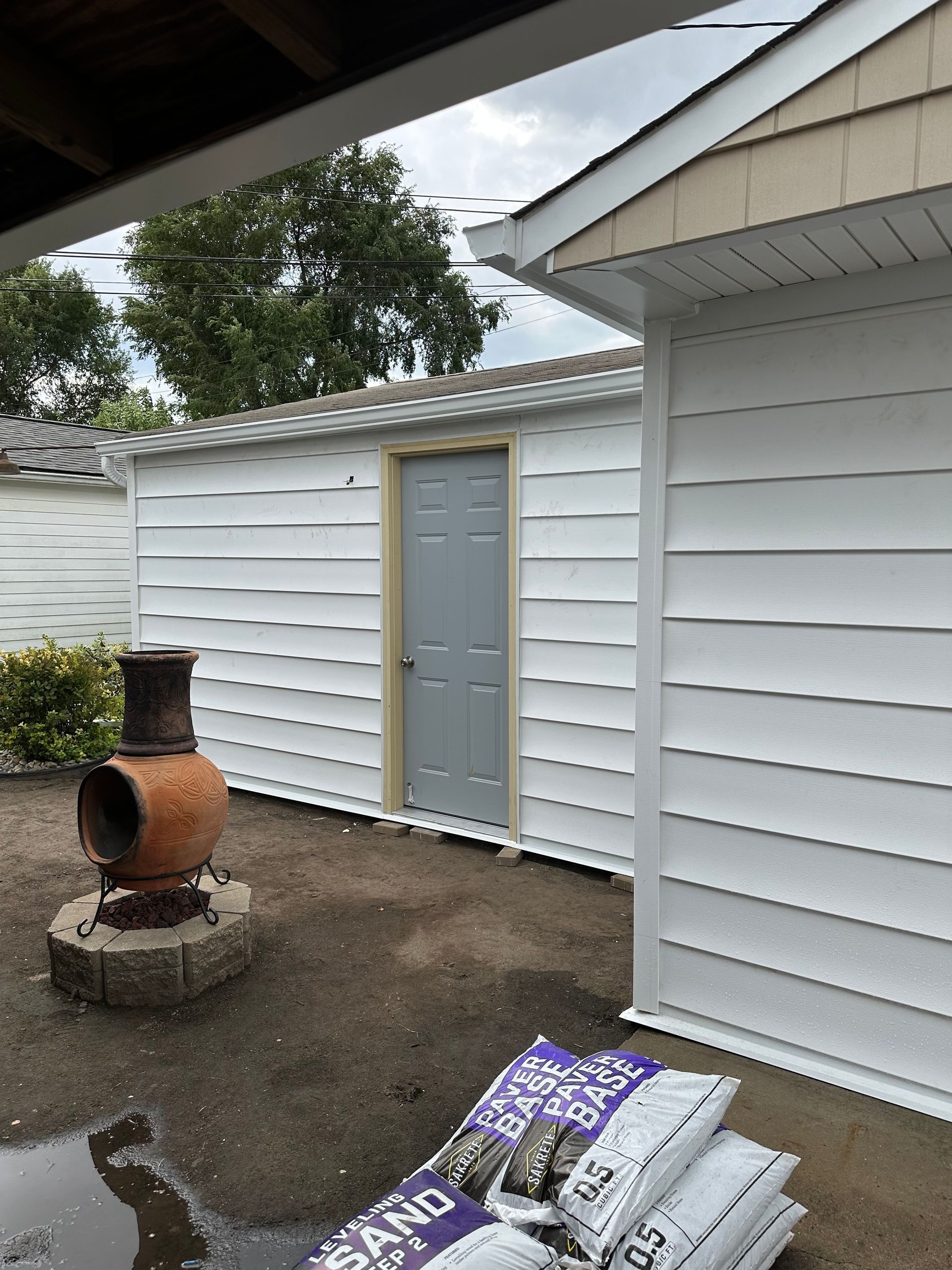 A white-sided shed with a gray door, next to a building with beige siding. Sandbags sit nearby.