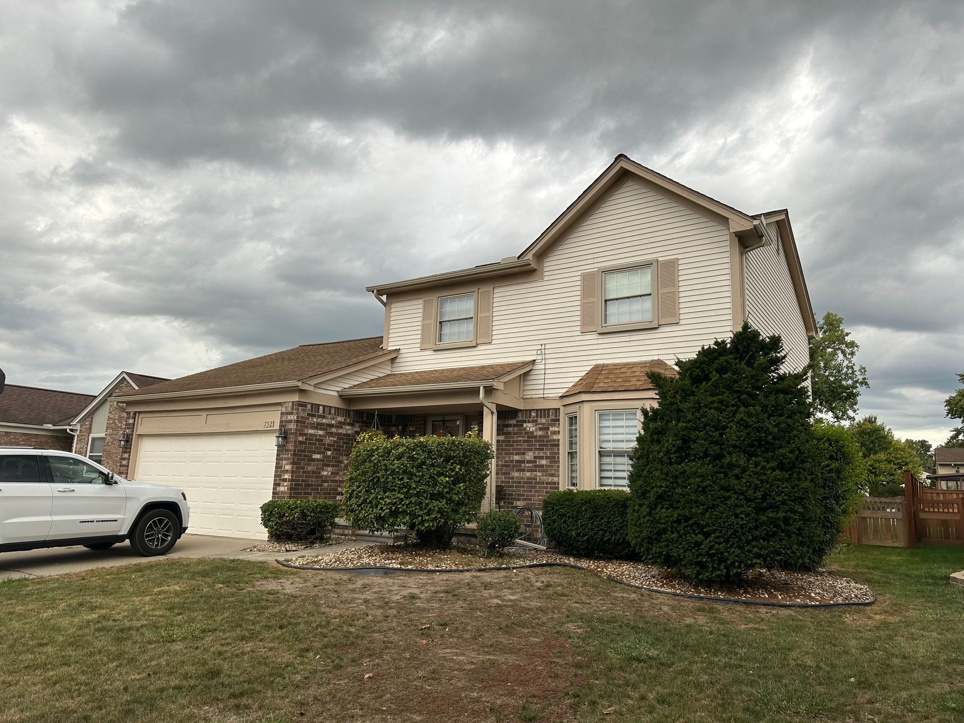 Two-story house with beige siding, brick accents, and a white garage door under a cloudy sky. A white SUV is parked in the driveway.