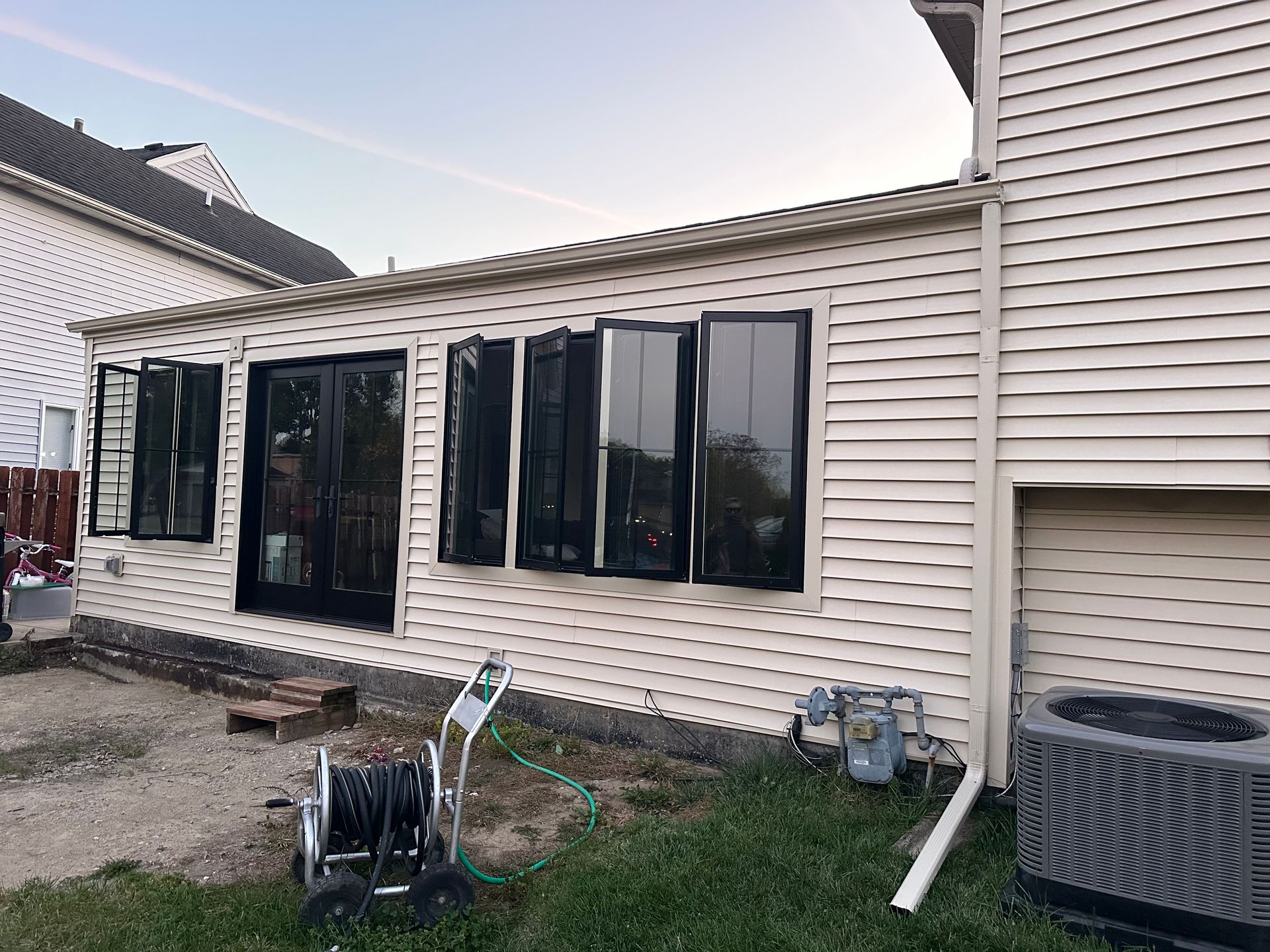 Exterior of a house with cream siding and black-framed windows. A hose reel sits on the ground.