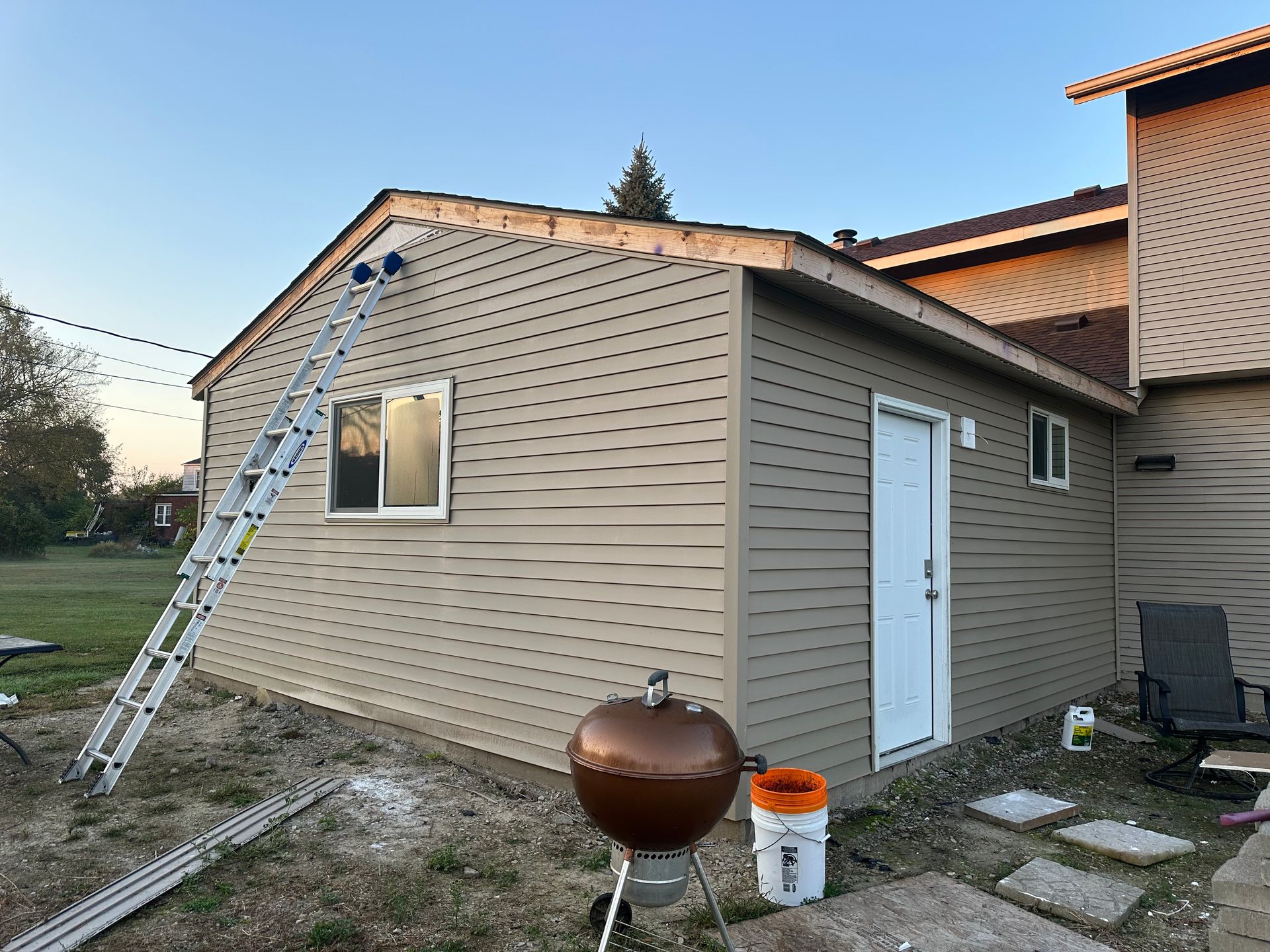 A building with beige siding under construction; a ladder leans against the wall. There's a grill and bucket nearby.