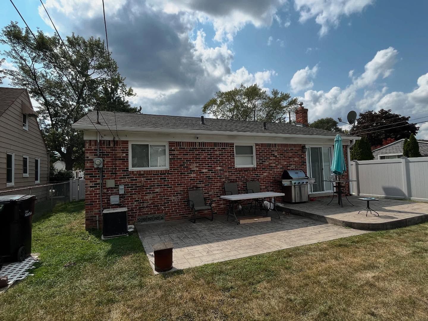 Backyard patio with brick wall, grill, table, and chairs under a cloudy blue sky.
