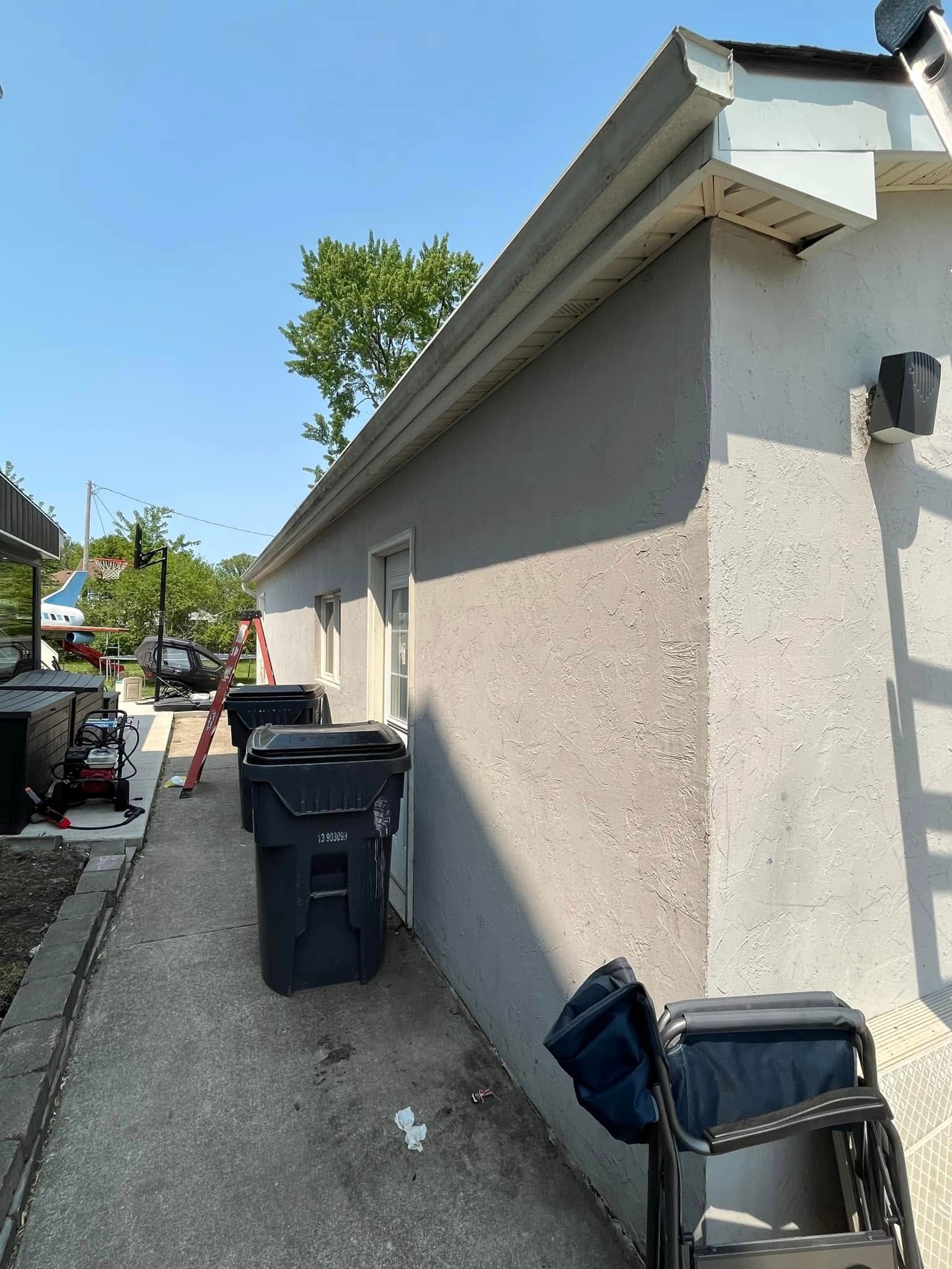 White house with black trim, overgrown yard. Trash bin on the right. Blue sky.