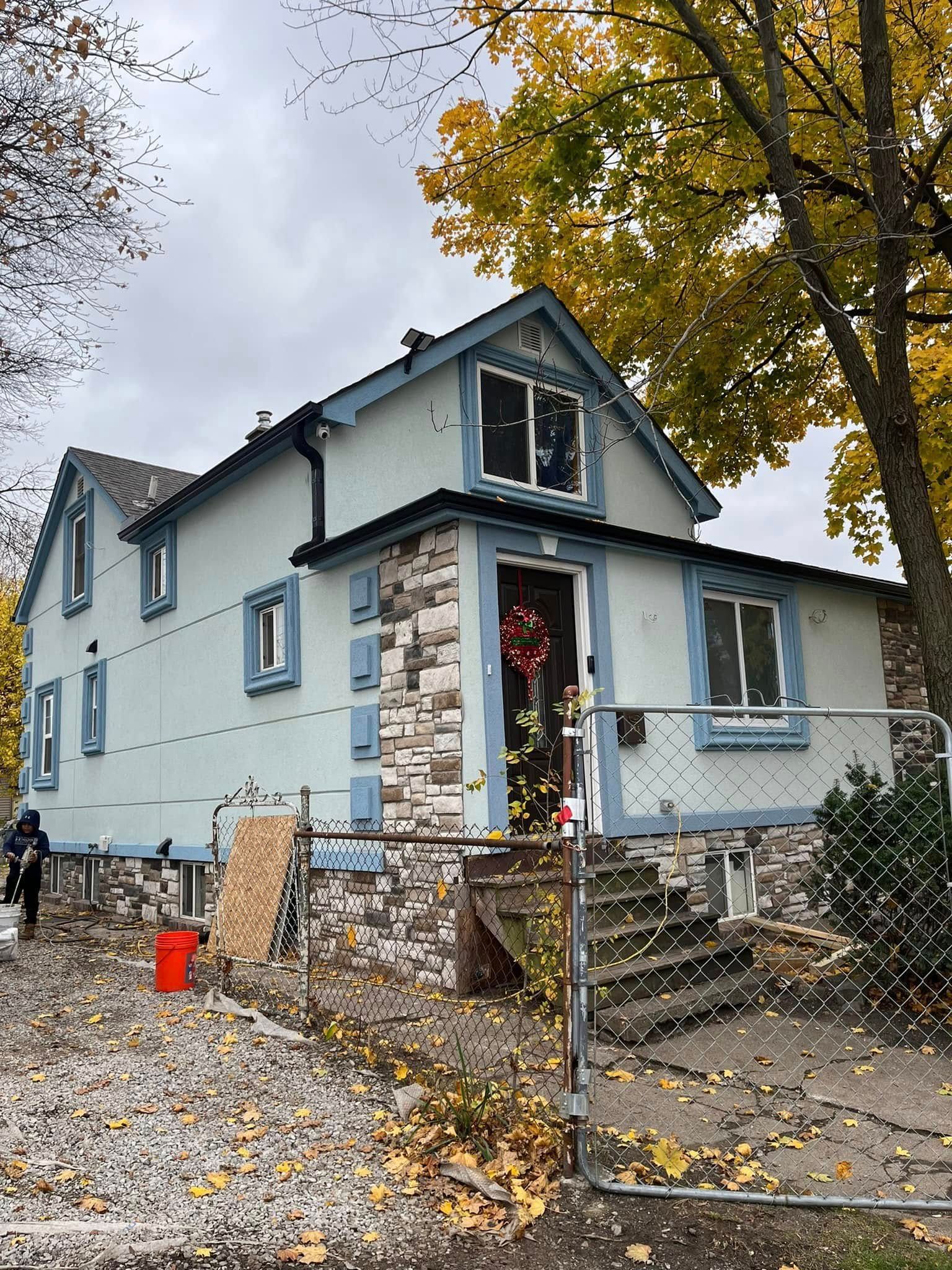 Two-story house with light blue siding, stone foundation, and a small porch. A person stands near the house.