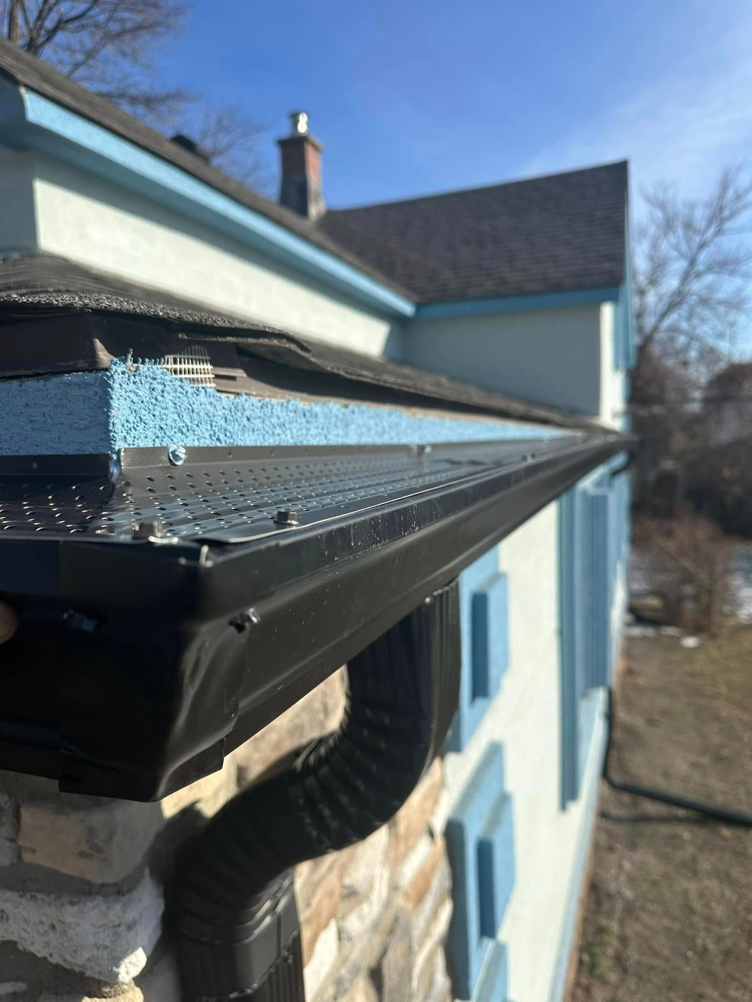 Black gutter and downspout on a light-colored house with blue trim; blue gutter guard visible.