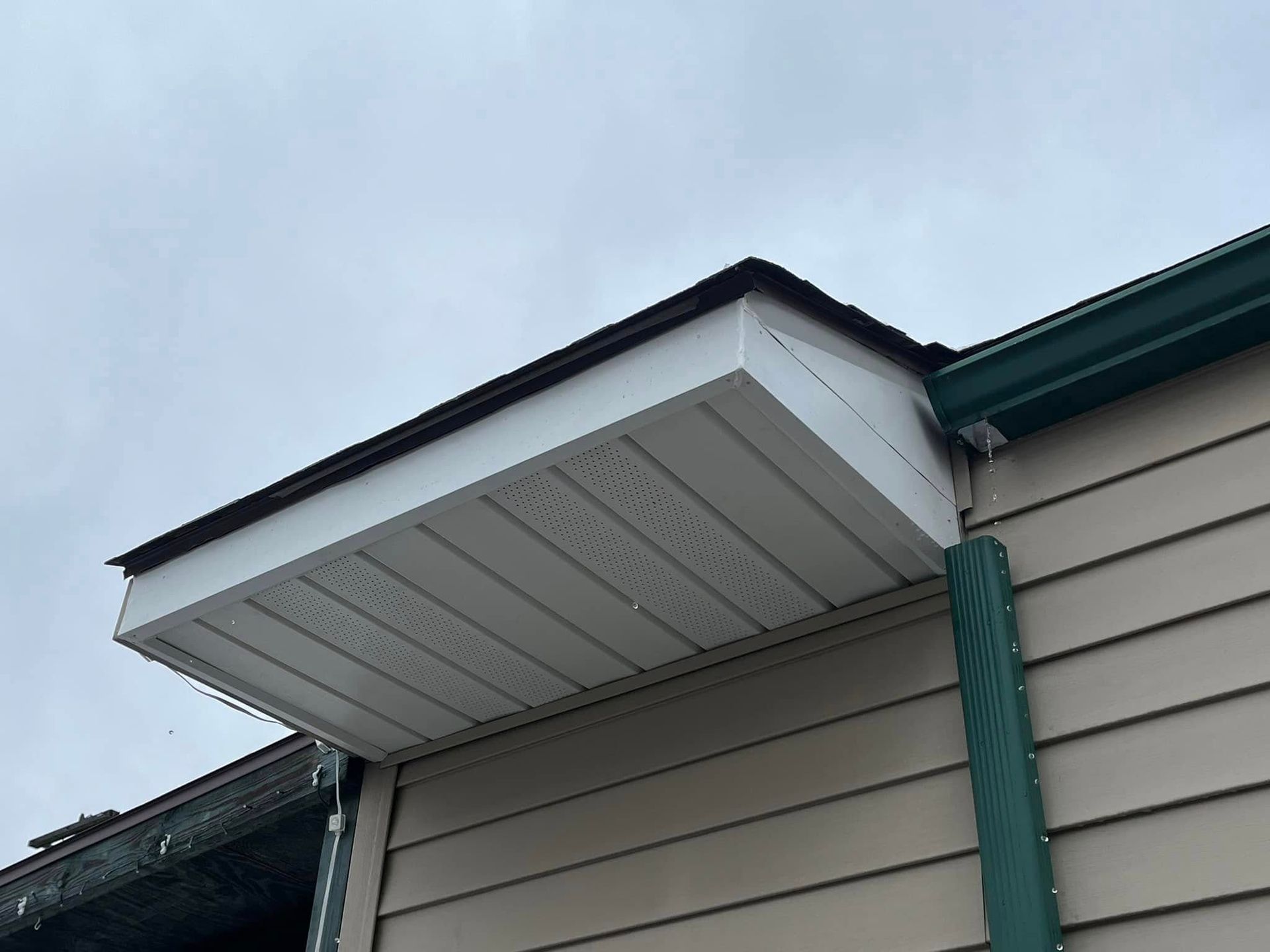 White soffit with brown trim on a tan house with green gutter under a cloudy sky.