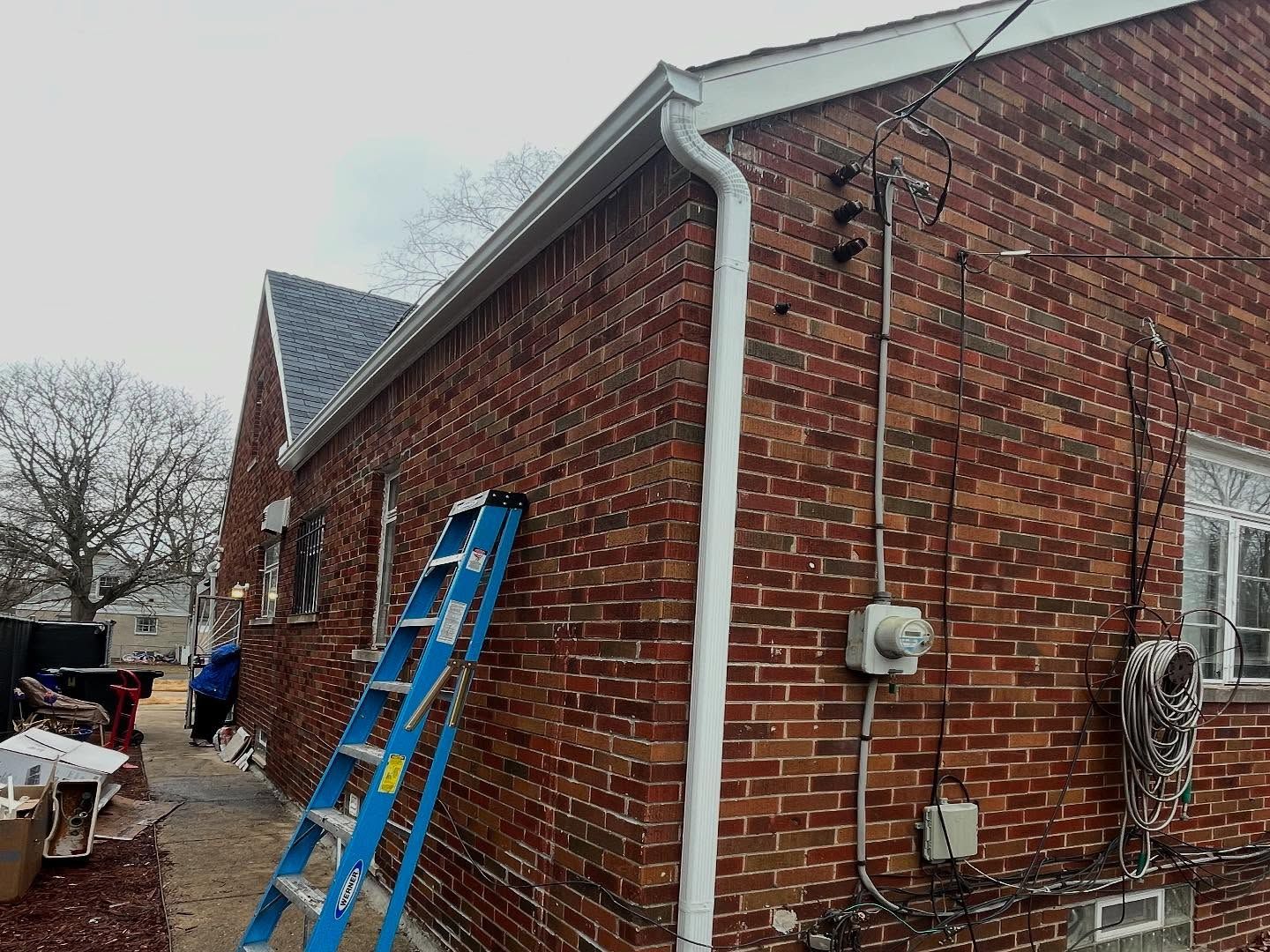 Red brick house with a blue ladder, white gutters, and electrical wiring on the wall.