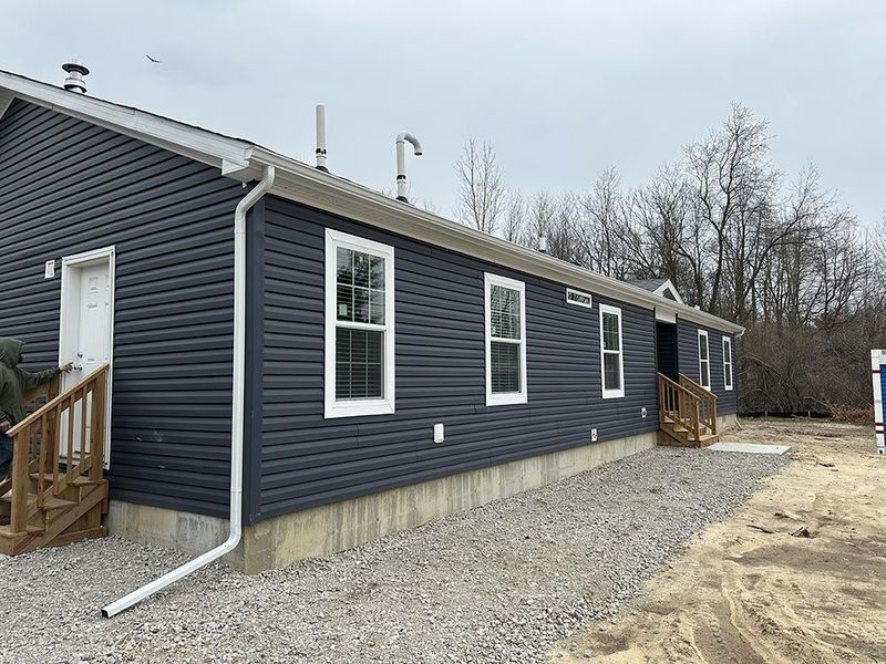 Dark blue sided building with white trim, windows, and rain gutters. Gravel ground.