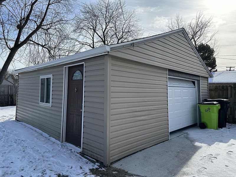 Tan sided garage with a white garage door, a brown door, and a small window in a snowy setting.