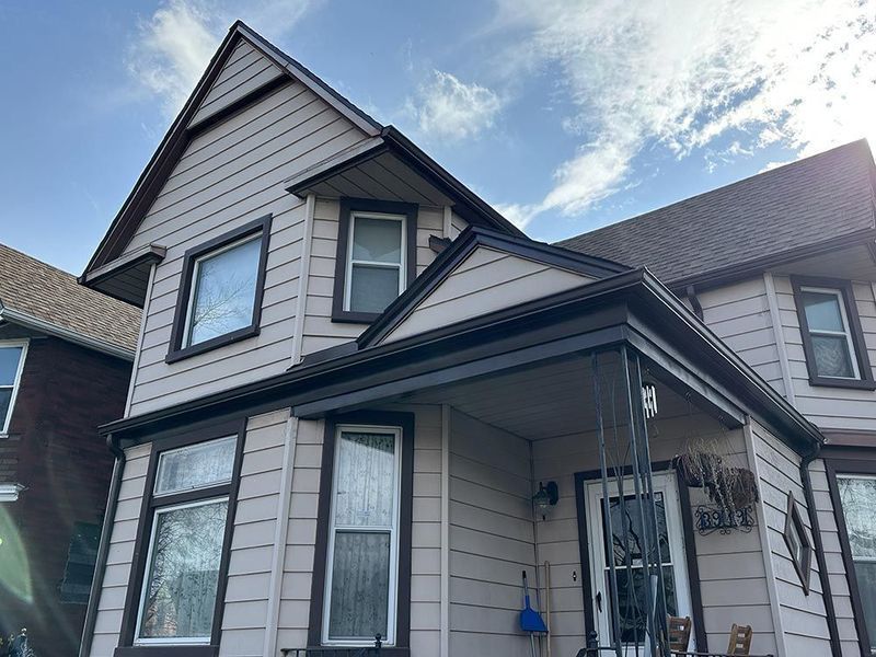 Two-story house with beige siding, dark trim, and a porch. Blue sky with clouds in the background.