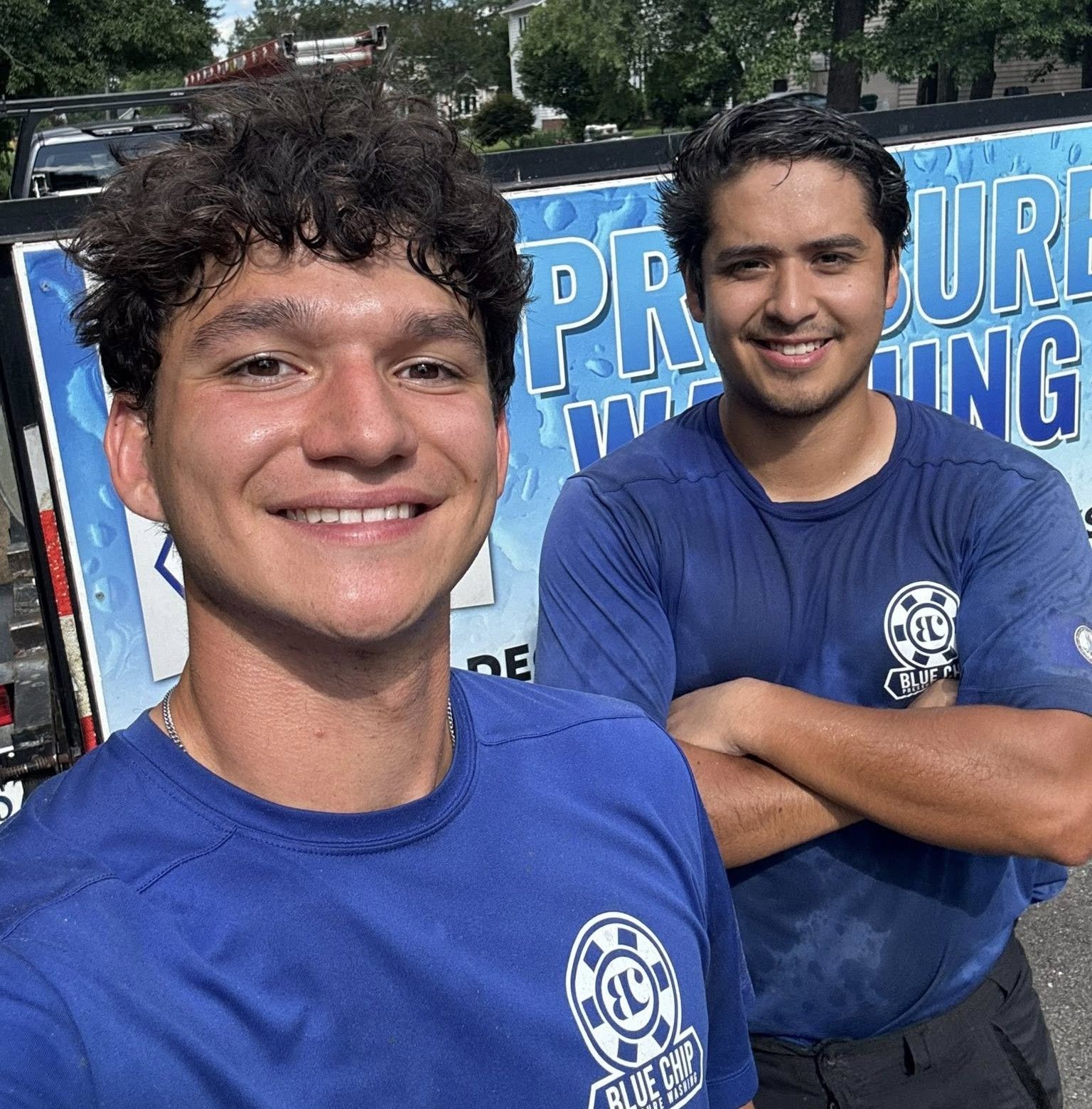 Two men in blue shirts smiling, standing next to a pressure washing sign.