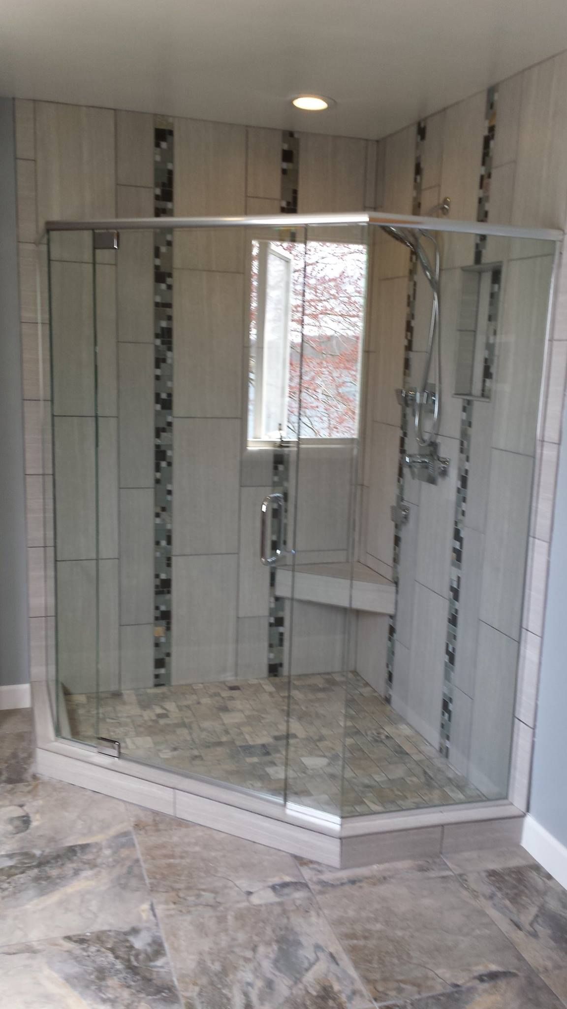 Glass shower enclosure with gray tile walls and bench, in a bathroom with stone flooring.