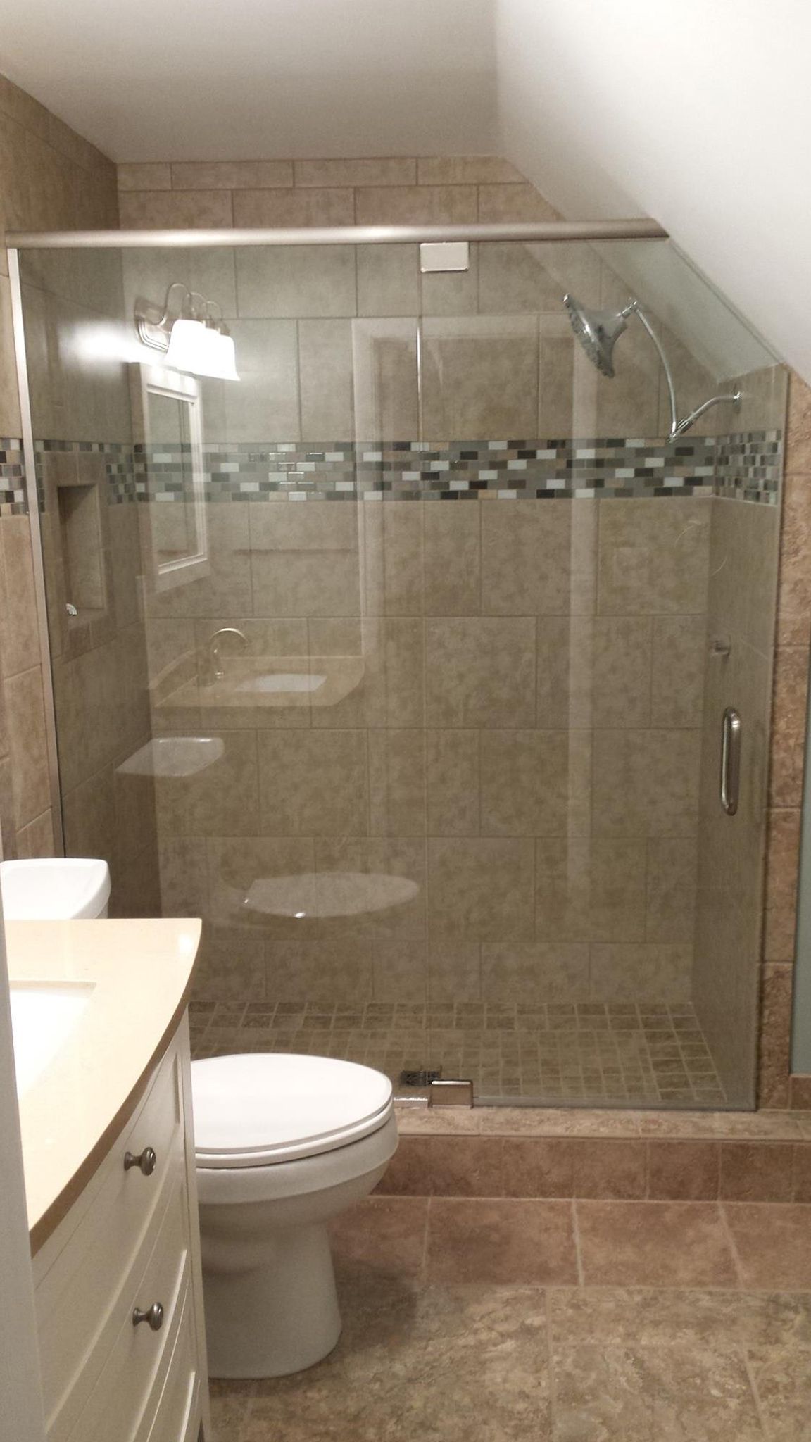 Bathroom with glass shower, toilet, vanity. Beige tile walls, decorative tile strip.