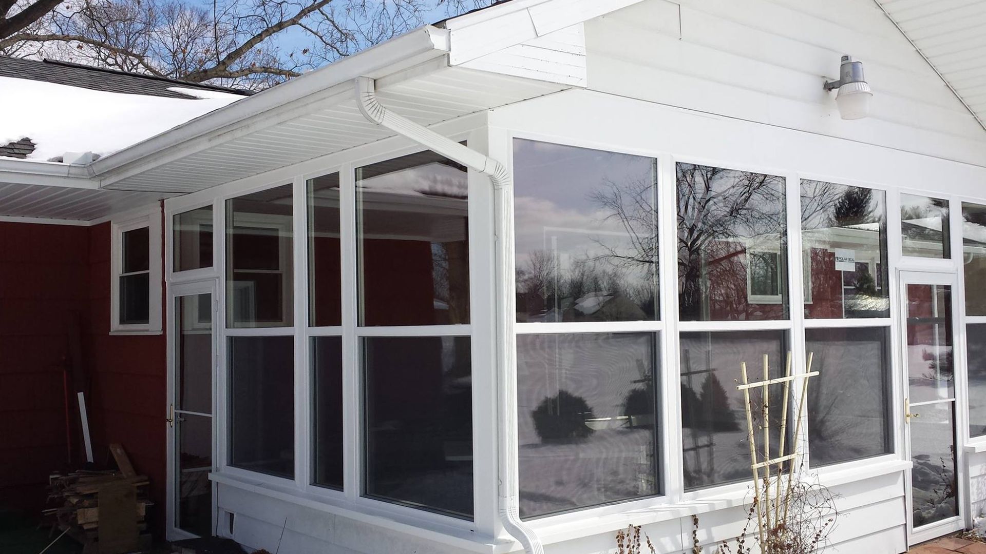 White-framed sunroom with many windows attached to a red house; snow visible on the roof.