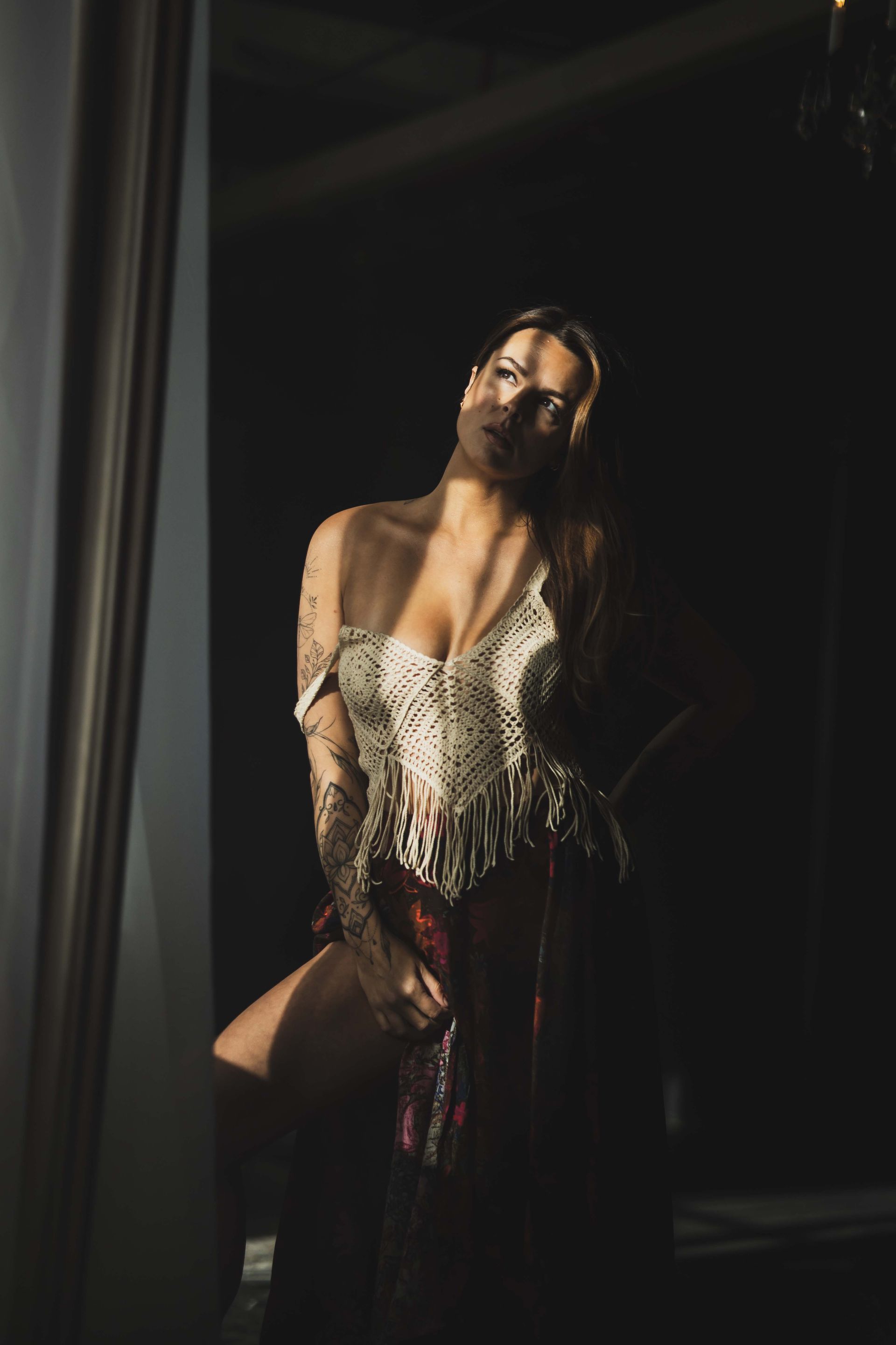 Woman in a dark room, lit by sunlight, wearing a sheer top, and holding up her skirt, looking up with a slight smile.