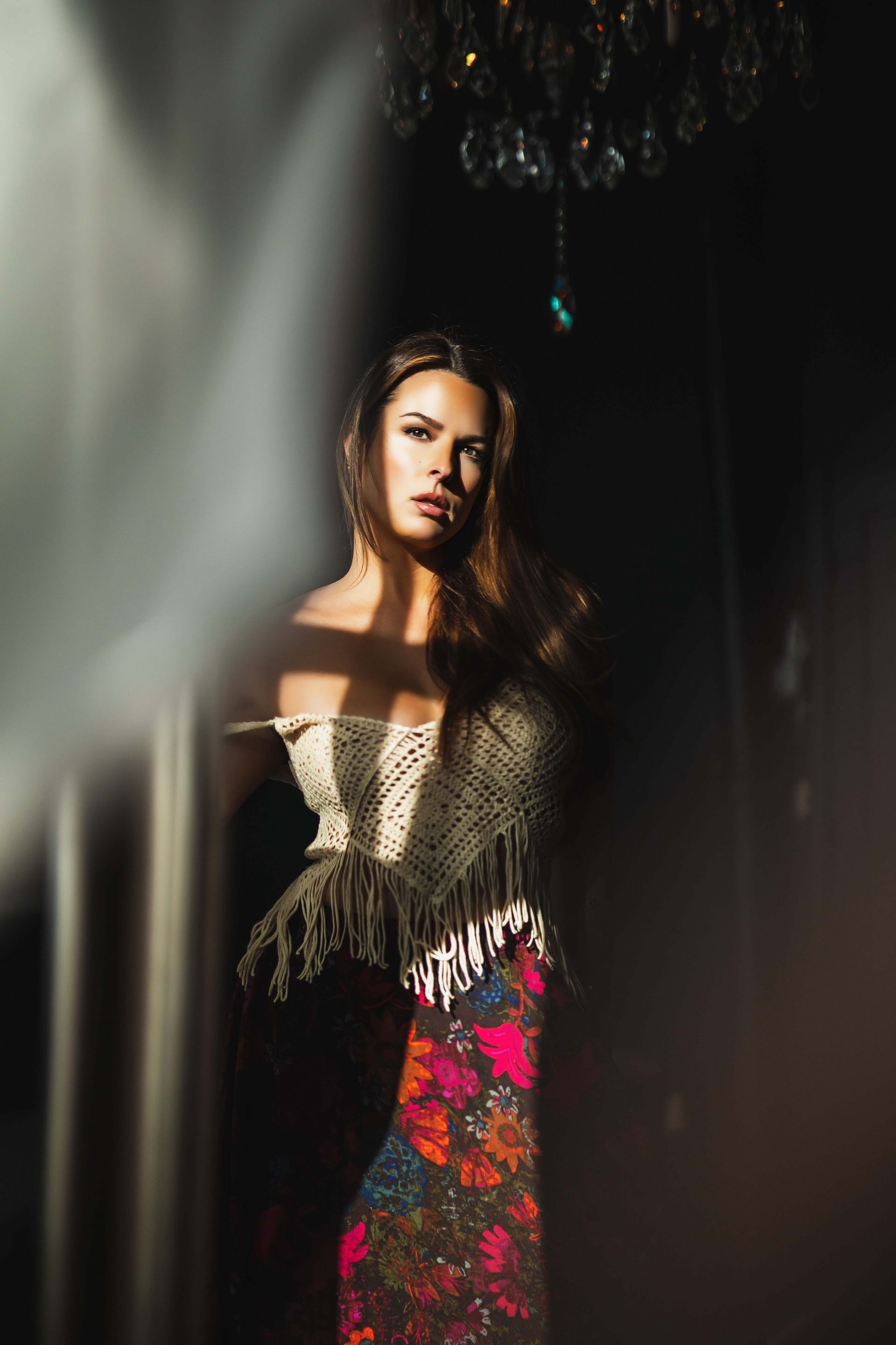 Woman in off-the-shoulder top and patterned skirt, lit by a shaft of light, stands in a dark room beneath a chandelier.