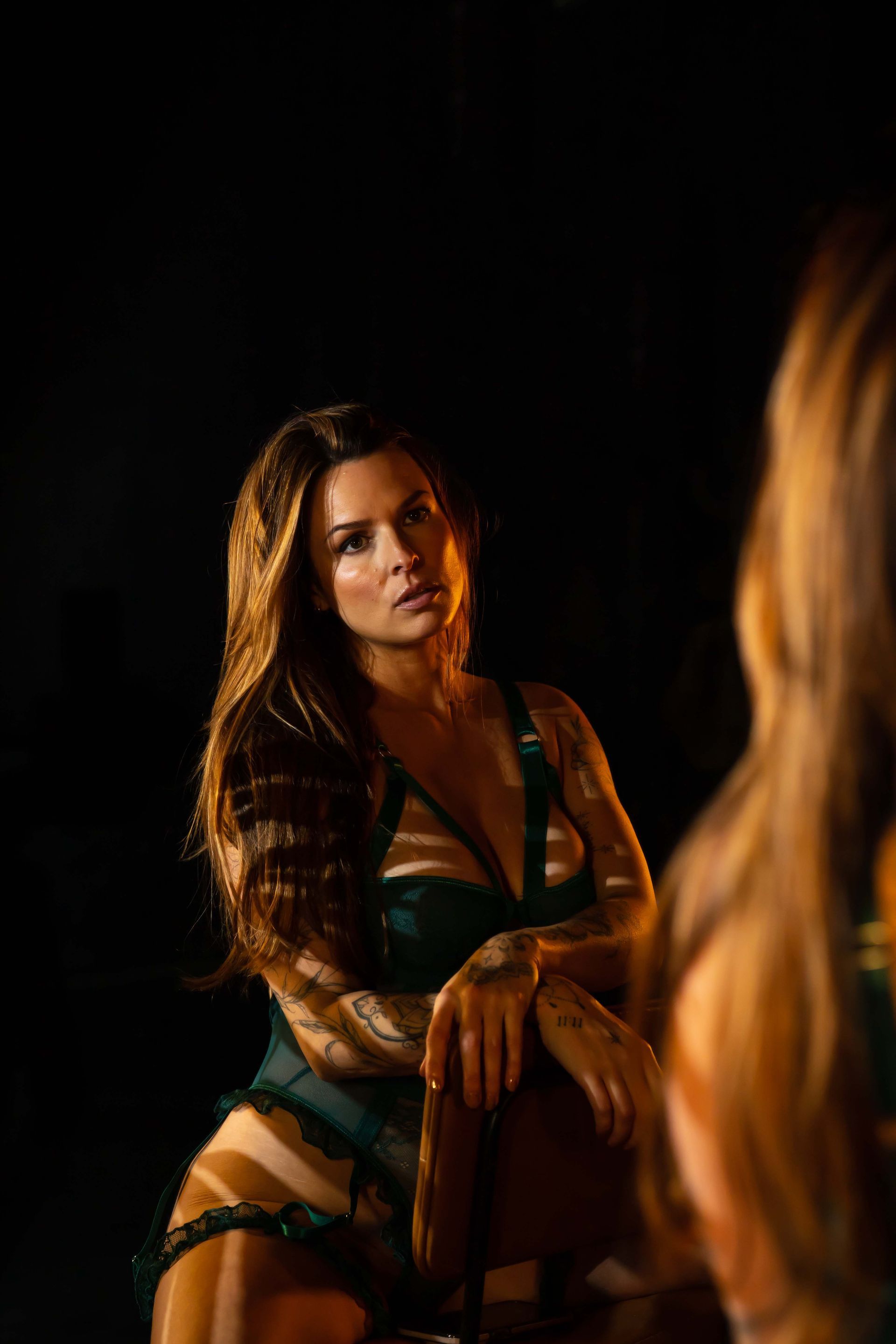 Woman in emerald lingerie poses by a mirror in a dimly lit room. Her reflection is seen.