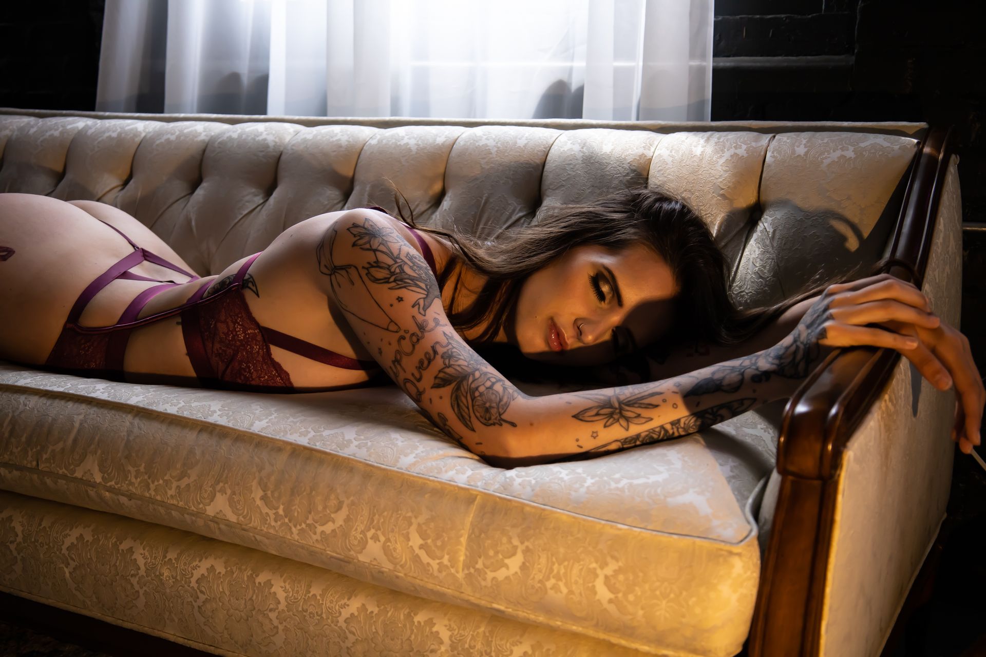Woman with tattoos in purple lingerie reclines on a white couch, looking at the viewer in a room with a window.