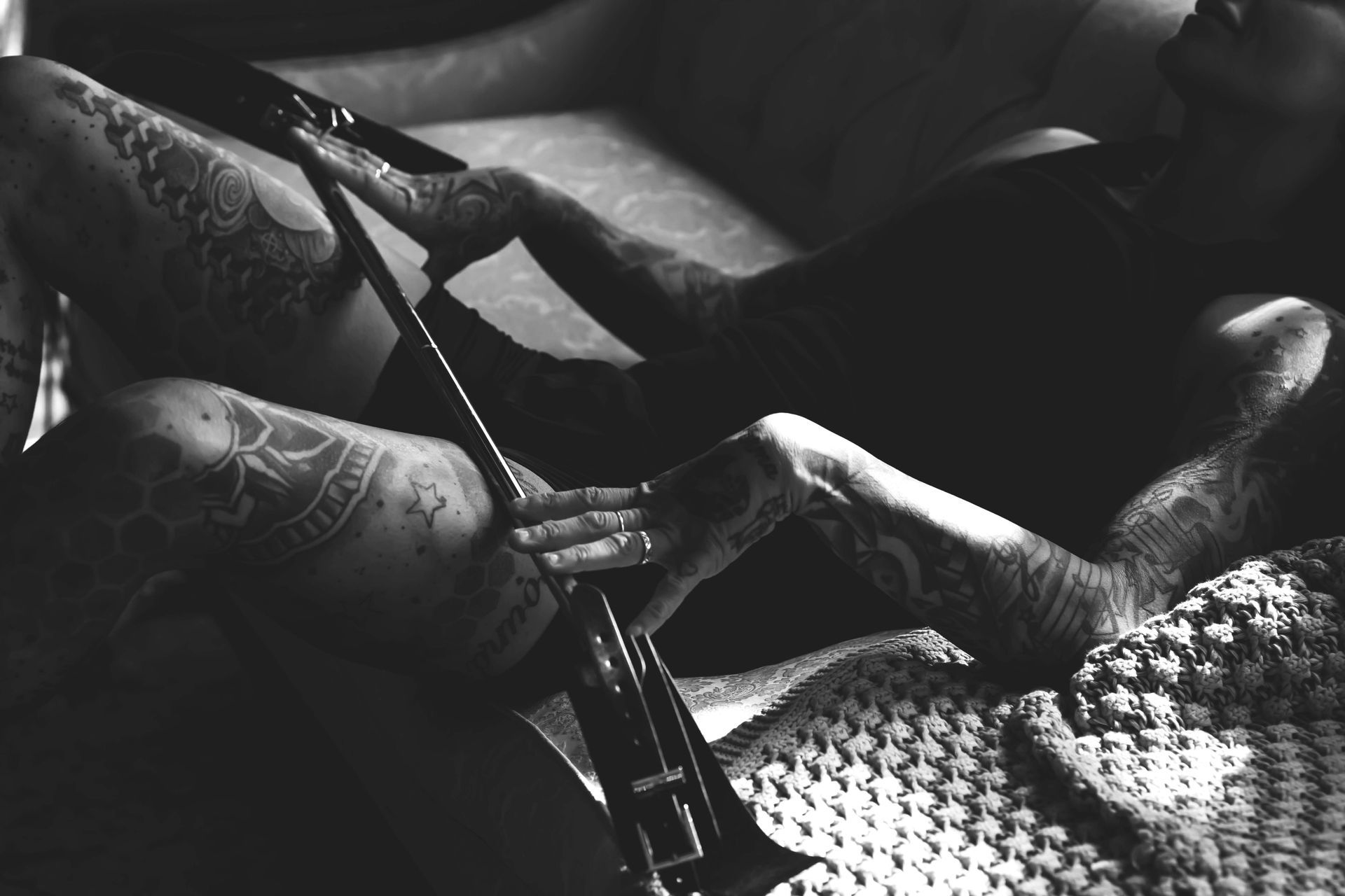 Black and white photo of a person with tattoos on their arms and legs holding a trumpet while reclining on a couch.
