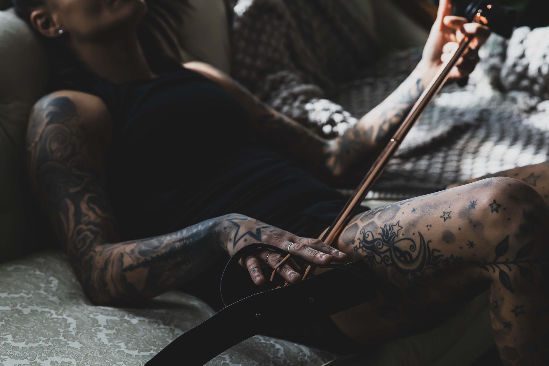 Person with tattoos playing a violin, resting on a couch. Warm sunlight, dark clothing.