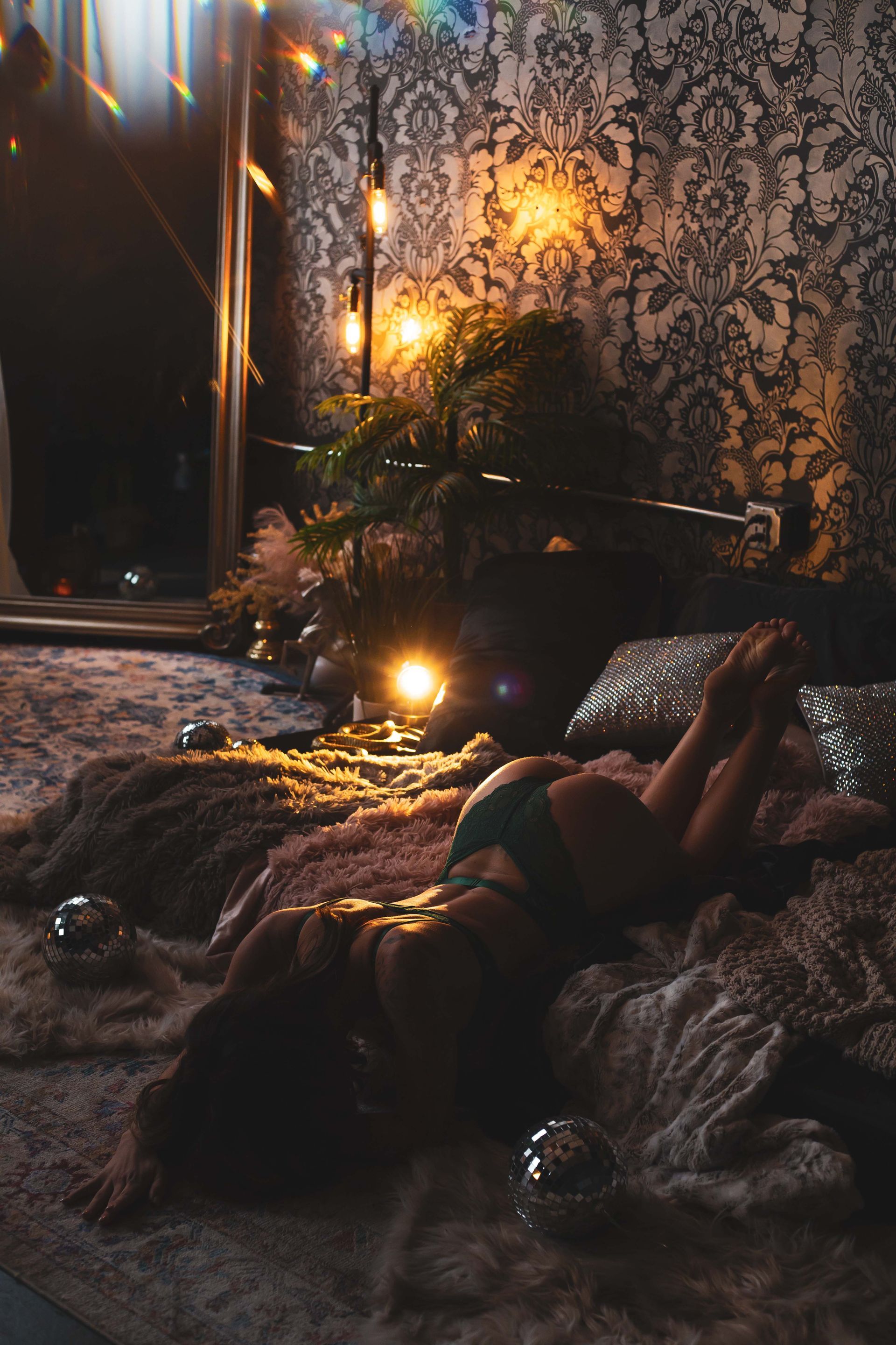 Woman in teal lingerie reclines on a fur rug, lit by soft lamps. A mirror and patterned wallpaper are in the background.