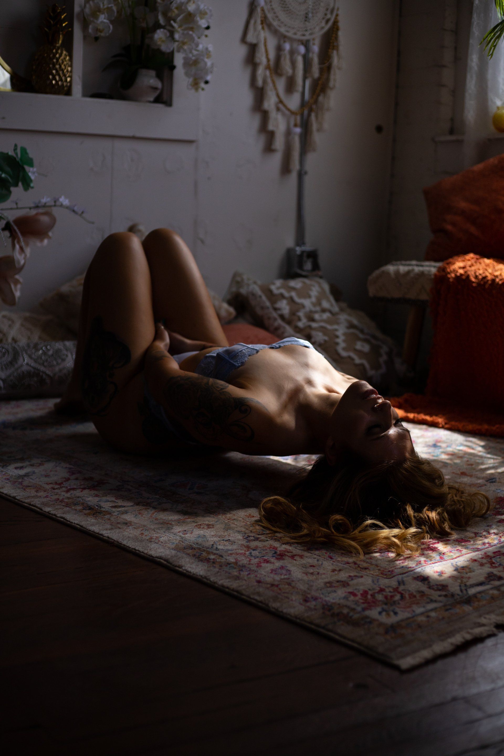 Woman lies on a rug in a sunlit room, head tilted back, arms raised. Sunlight streams in.