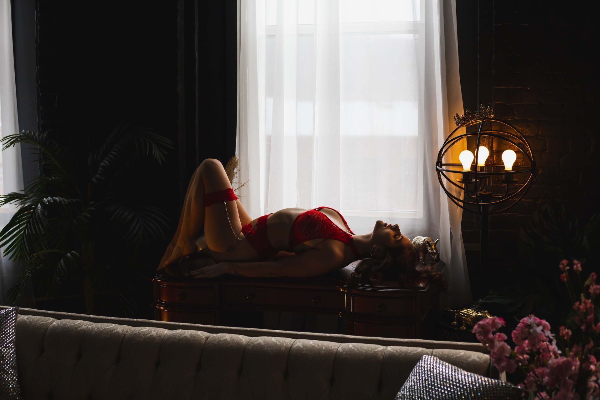 Woman in red lingerie reclines on a bench by a window. The room is dimly lit with a decorative light fixture.