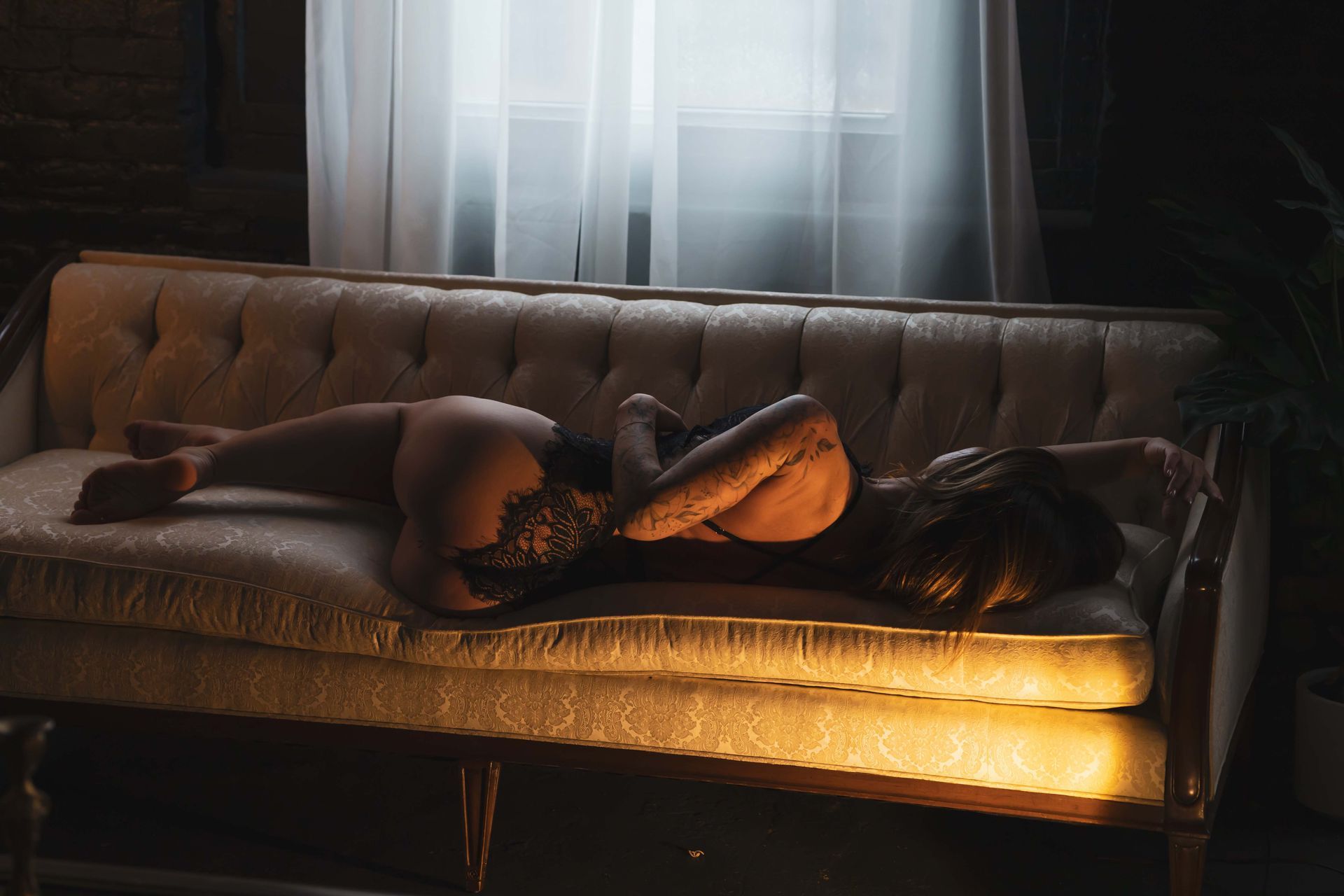 Woman in lingerie lies on a cream-colored couch, back to the viewer, in a dimly lit room. Light shines in from a window.
